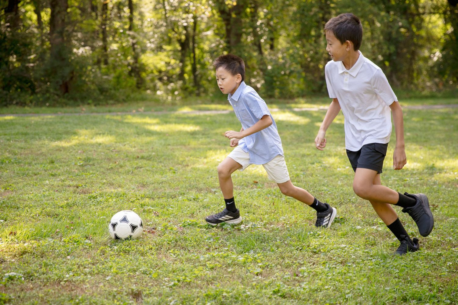 Two young boys playing soccer on a green grass field with trees in the background on a sunny day.