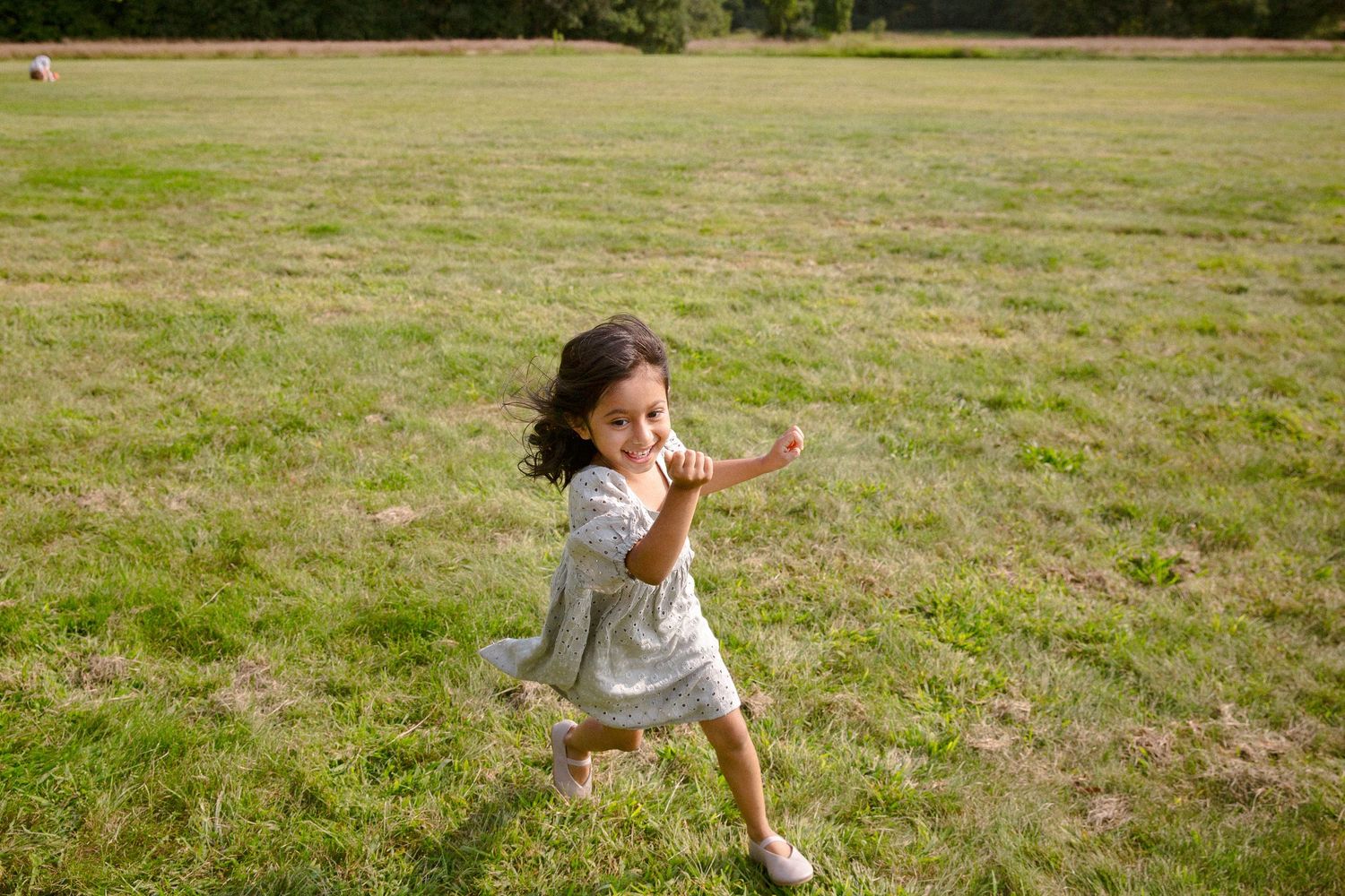 Carefree little girl in gray dress running barefoot across green grass field.