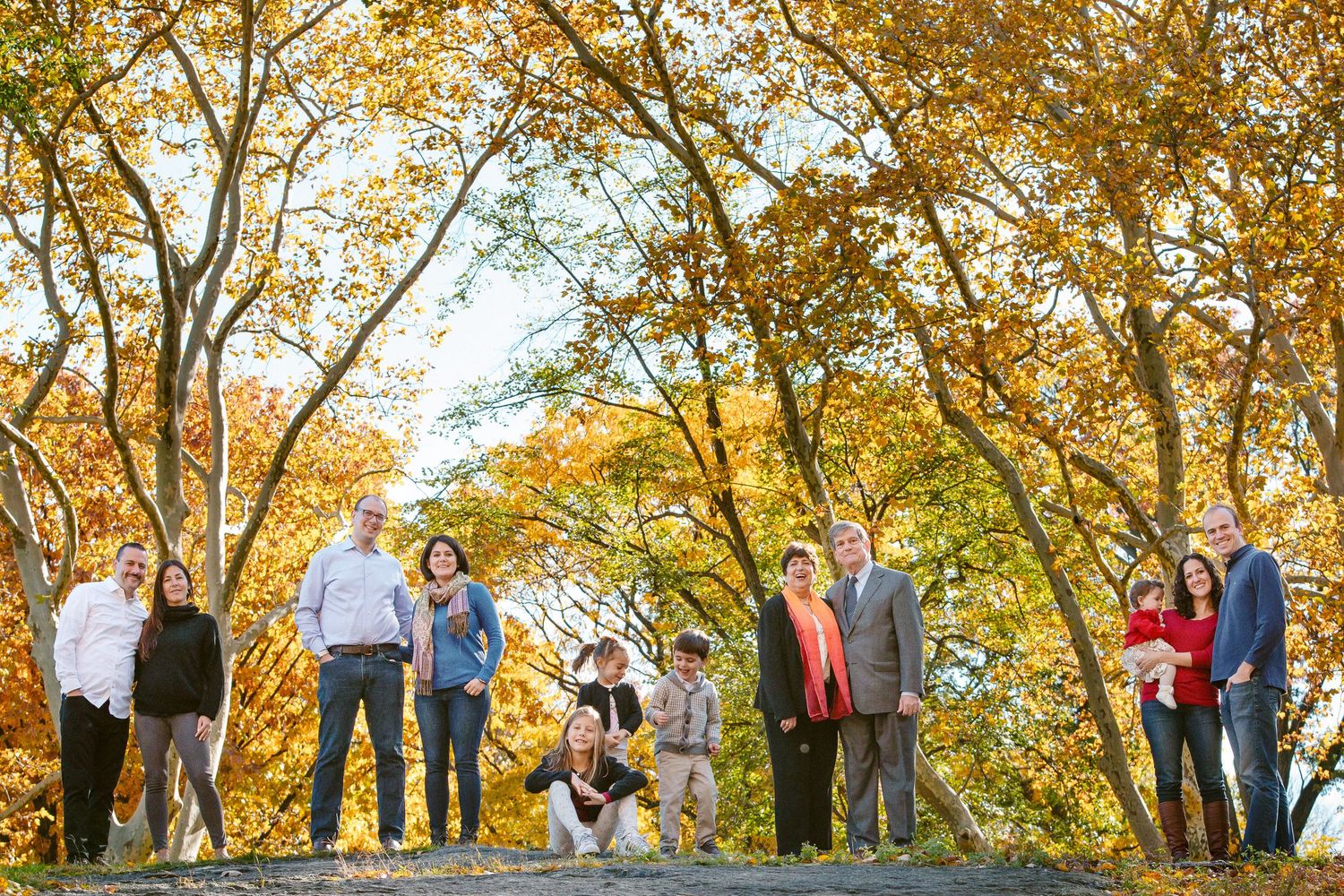 Large extended family posing together outdoors among vibrant autumn foliage trees in fall season.