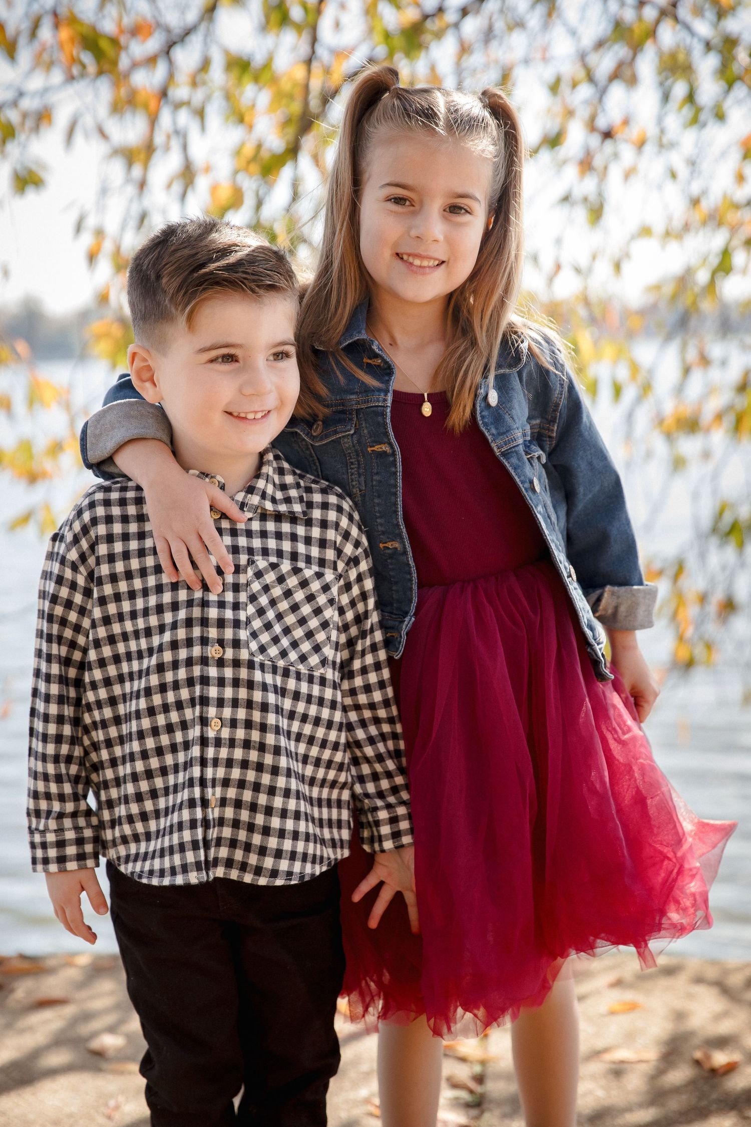 Young brother and sister pose together near a lake in autumn outfits.