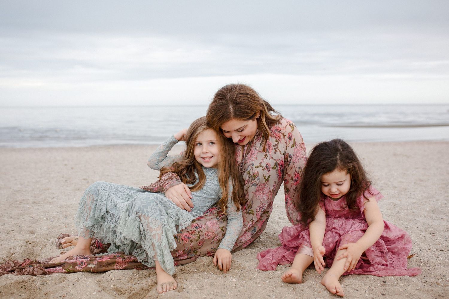 Mother and two daughters in floral dresses sitting together on sandy beach by ocean.