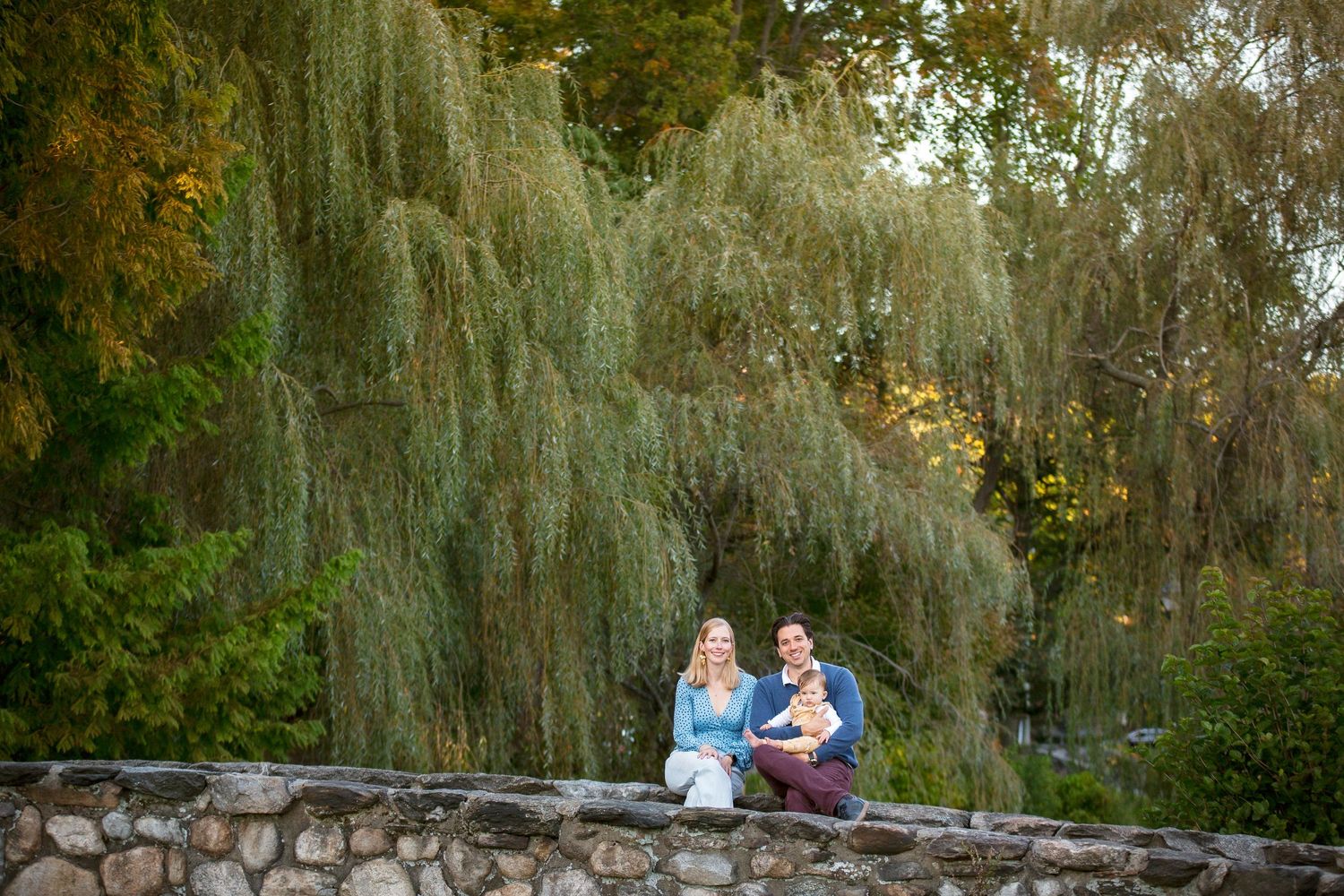 Young family with baby sitting on stone wall beneath weeping willow tree in park.
