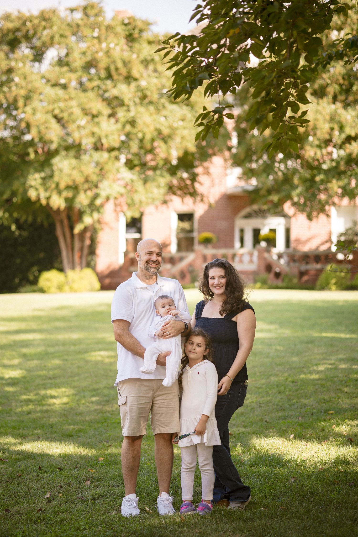 Family of four poses outdoors on green lawn with brick building and trees in background on sunny day.