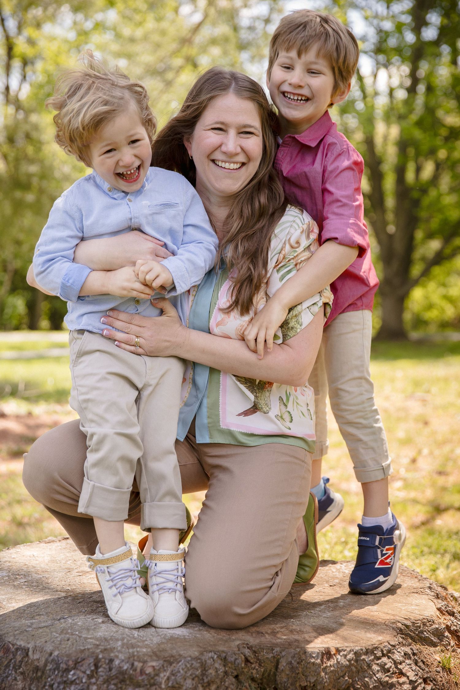 A joyful mother kneels on a tree stump hugging her two laughing young sons outdoors in a sunny green park.