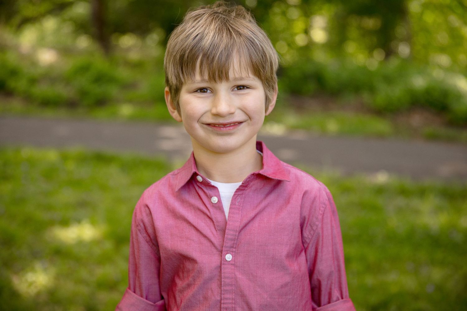 Smiling young boy with blonde hair wearing a red button-up shirt outdoors in a green park setting.