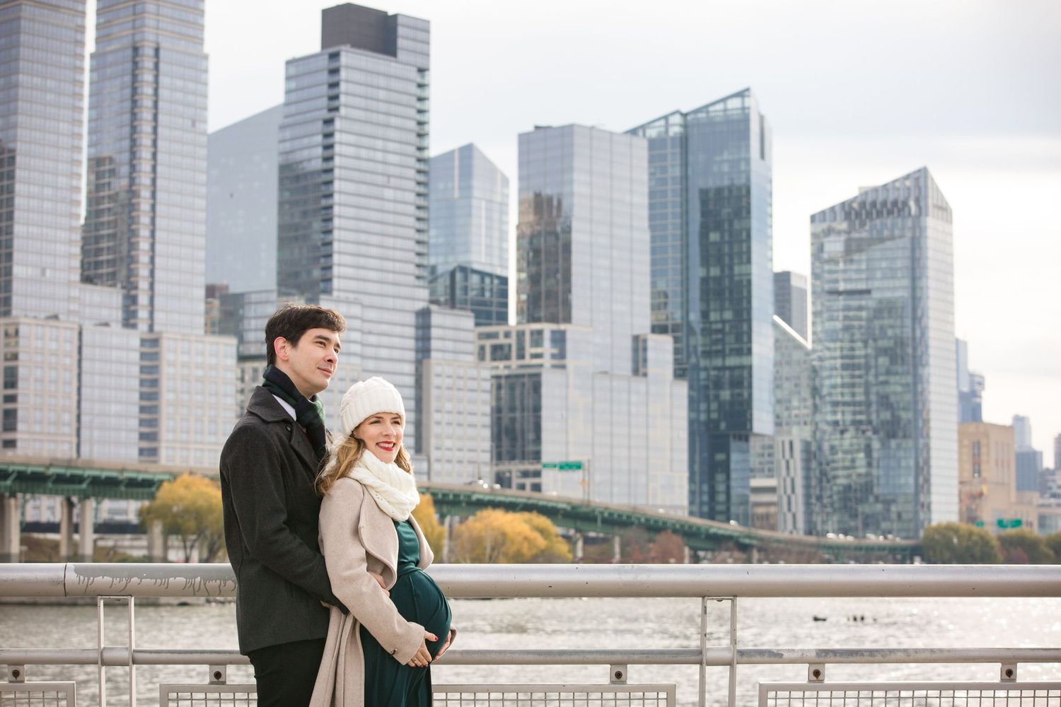 Expecting couple stands together by NYC waterfront with Manhattan skyline skyscrapers in background on cloudy autumn day.