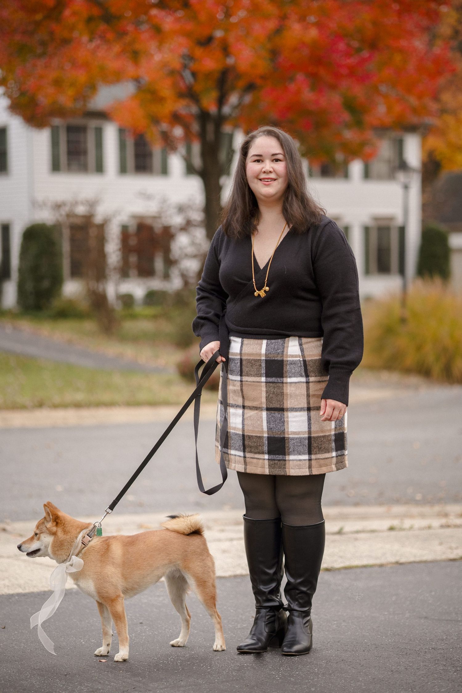 Woman in plaid skirt and black boots walking a Shiba Inu dog on a leash during autumn.