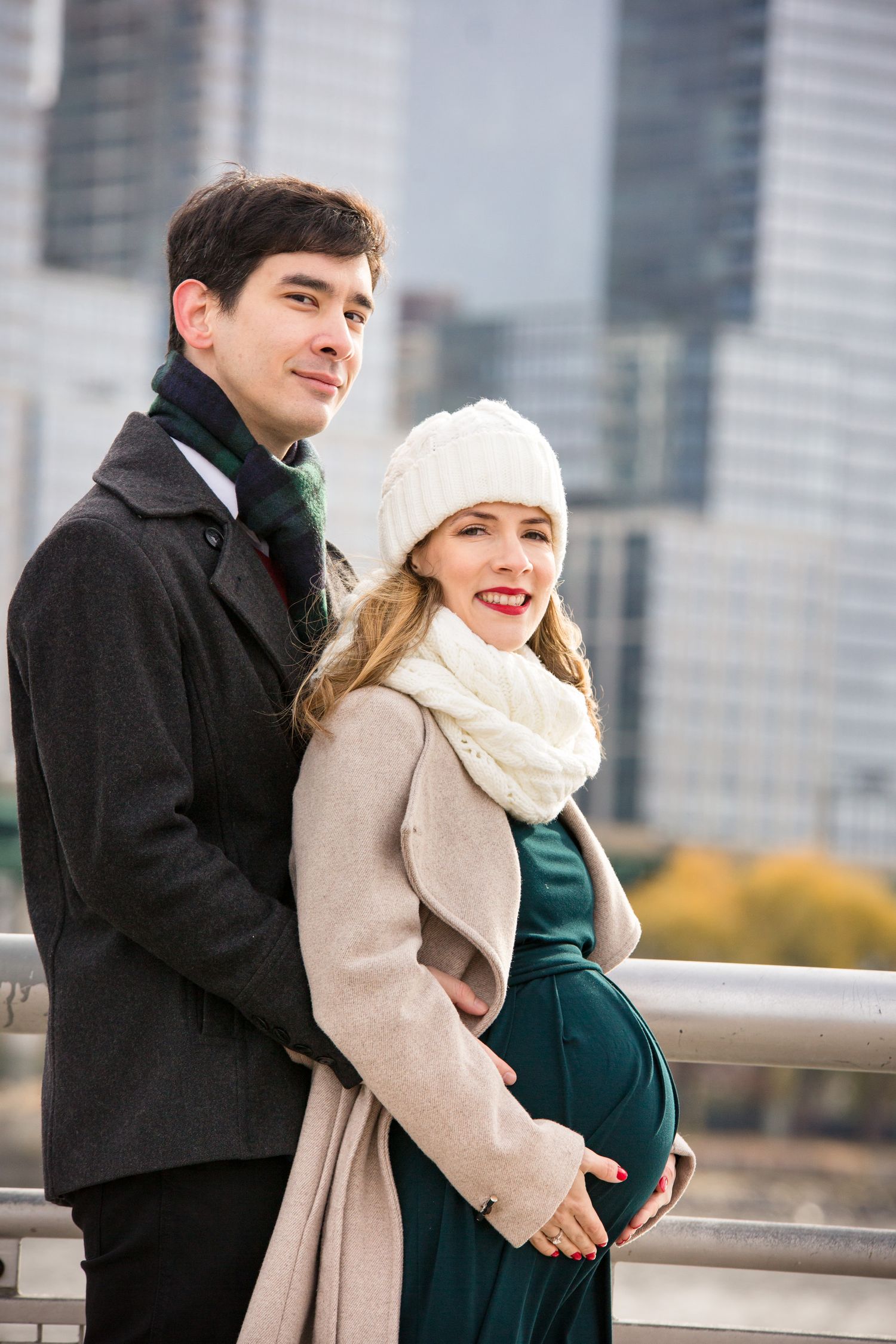 A happy couple poses outdoors in winter coats, the woman pregnant in a green dress and white hat.