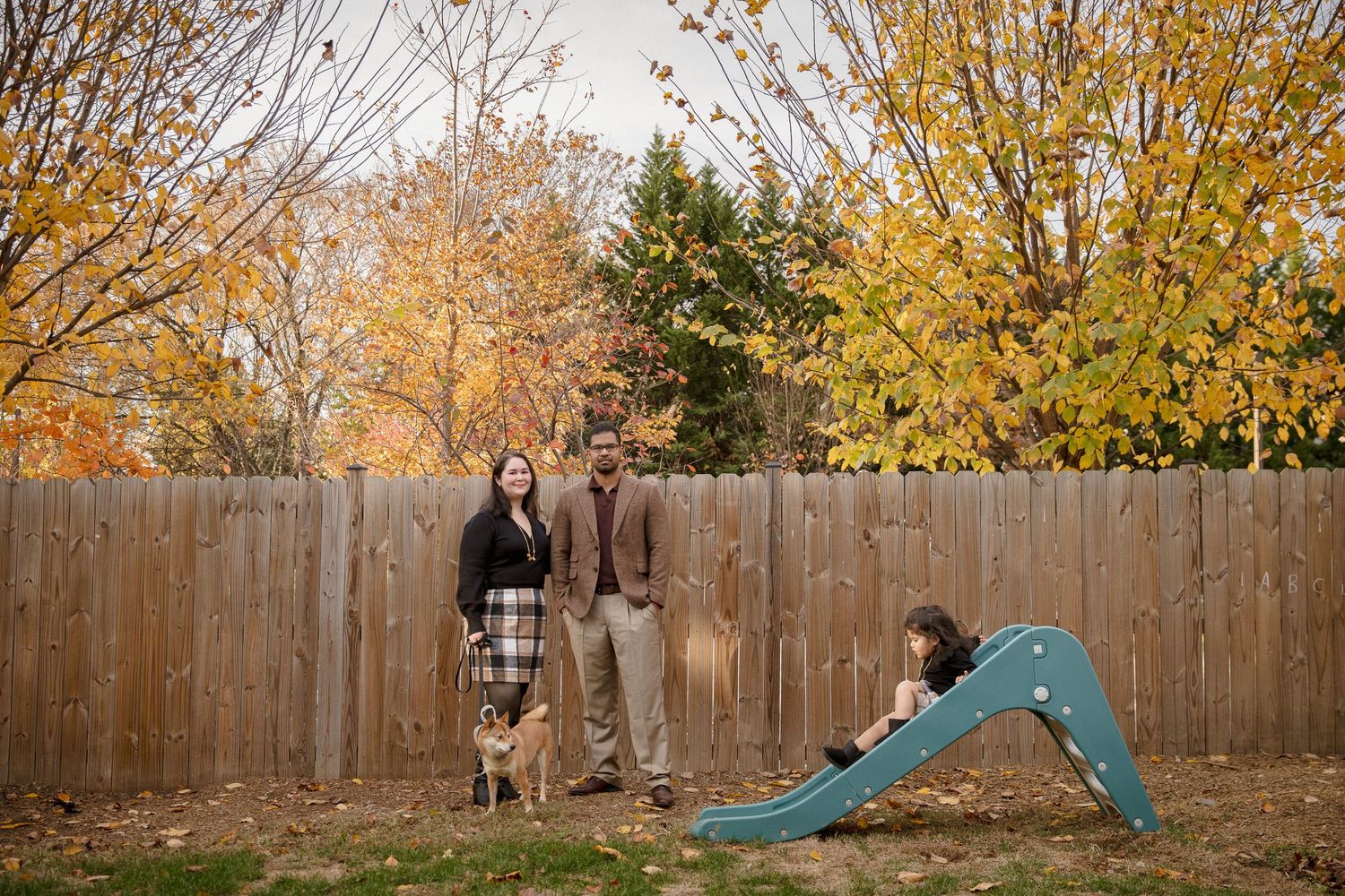 Family with dog and child on slide posing in backyard surrounded by colorful autumn foliage.