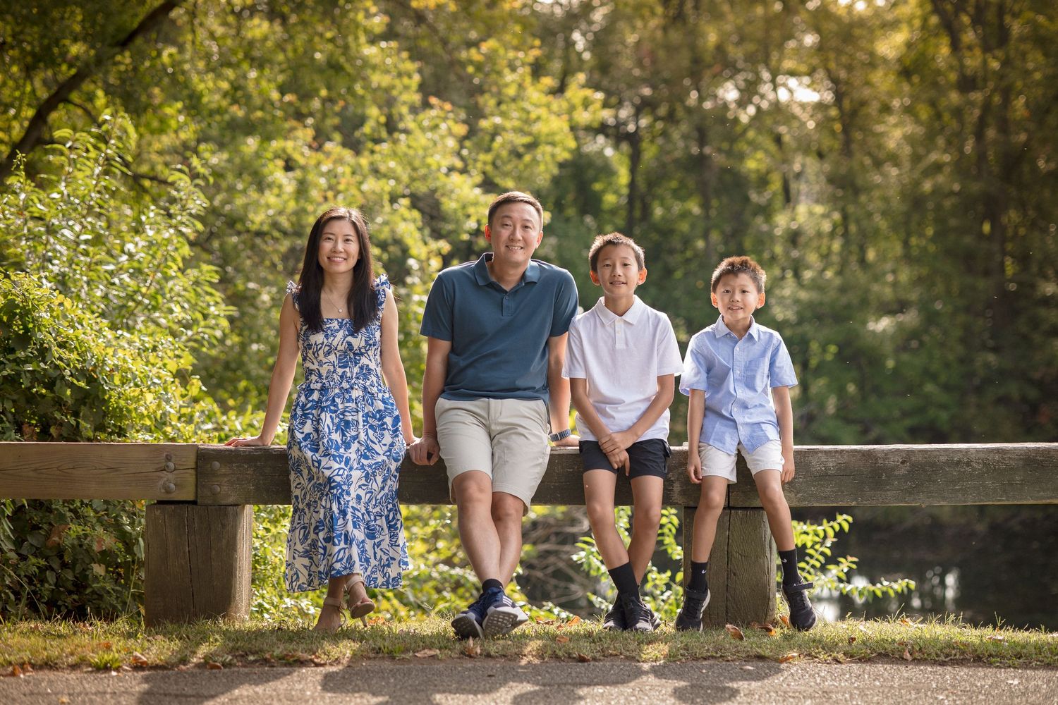 Family of four smiling outdoors on a sunny day, posing on a wooden bridge surrounded by lush green trees.