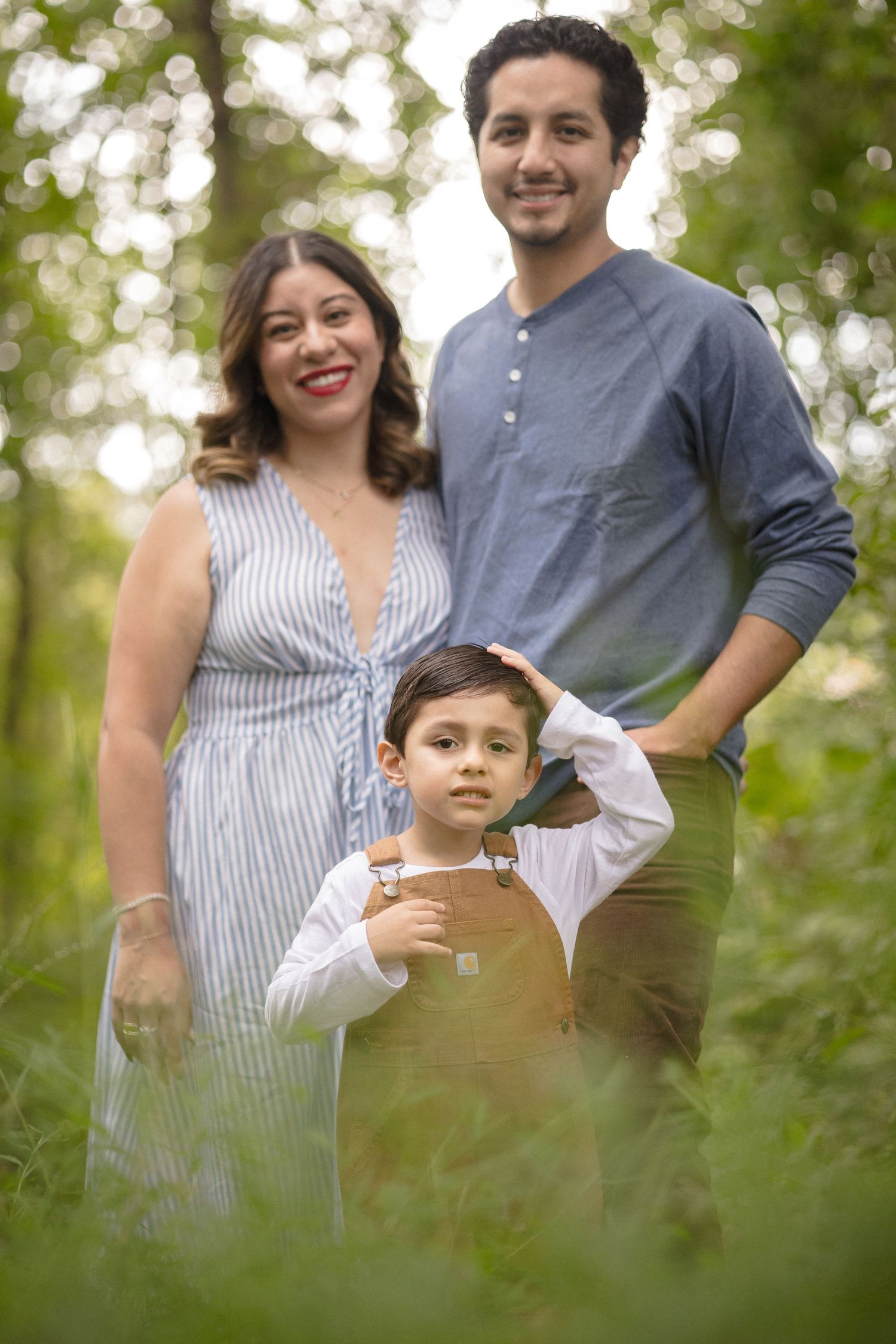 A smiling family of three poses together in a lush green forest setting.