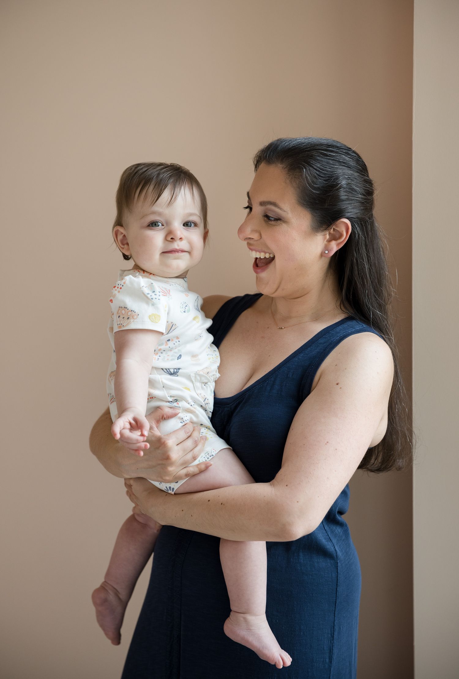 A smiling woman in a navy dress holds a happy baby in a white onesie against a beige background.