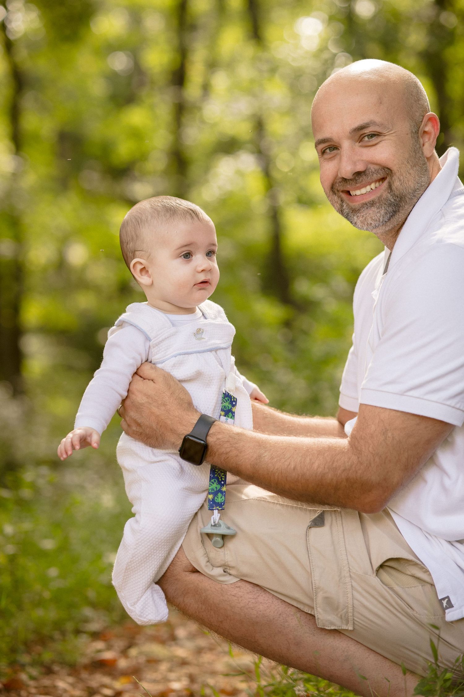 Smiling bald father holding adorable baby in white outfit outdoors in sunny green park setting.