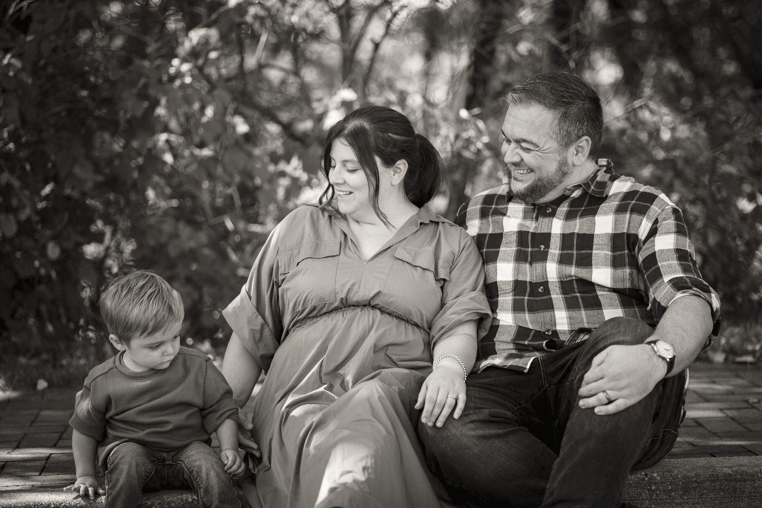 Black and white family portrait of parents and young child sitting outdoors, smiling together in a lush garden setting.