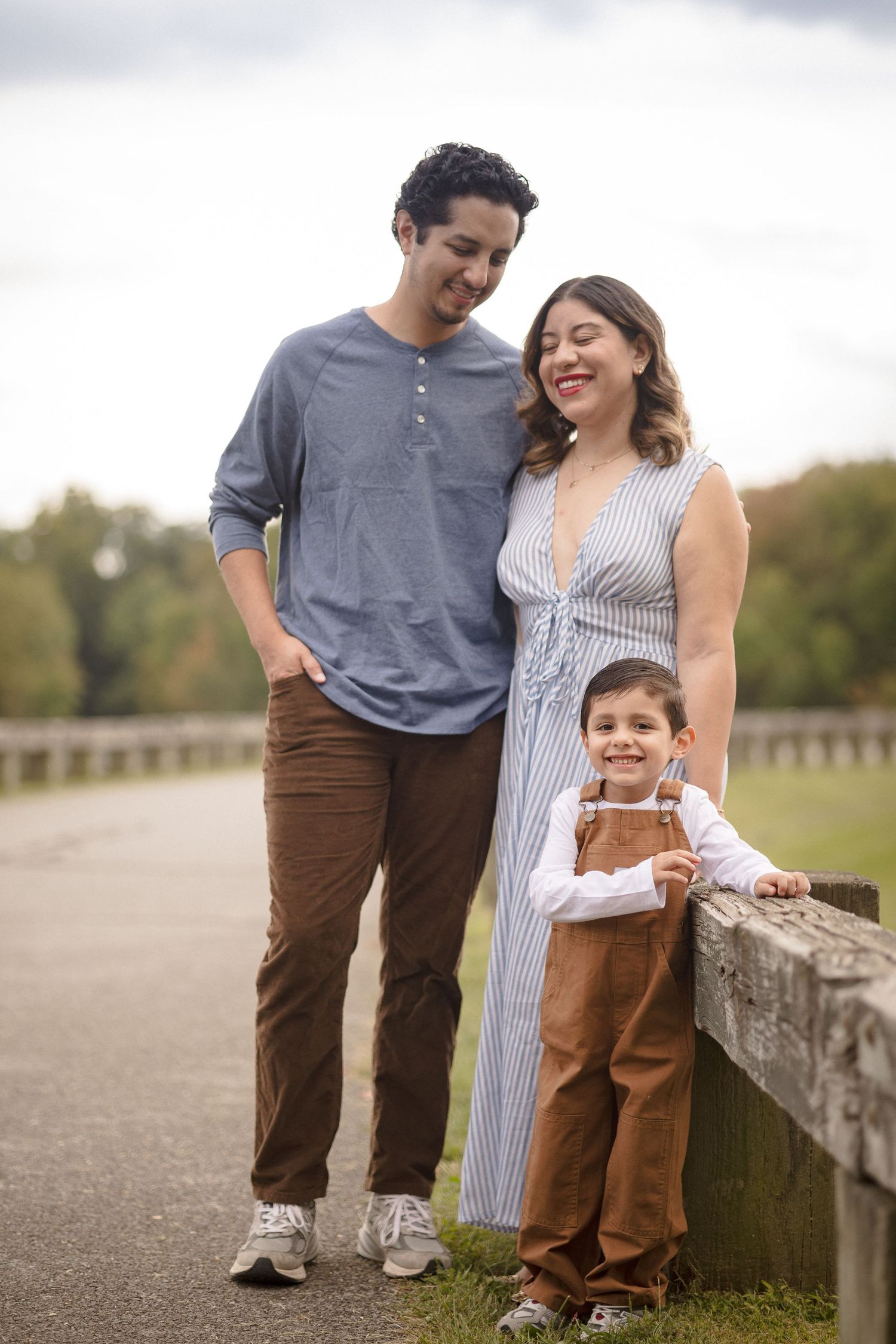 A happy family of three poses outdoors, with parents and young boy in overalls leaning on a wooden fence.