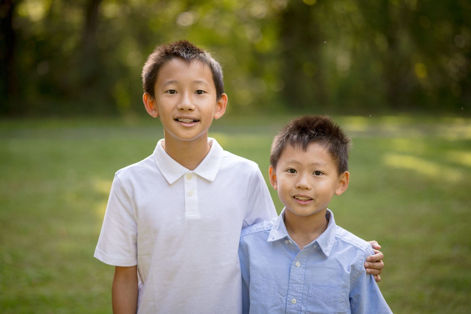 Two young Asian boys smiling outdoors in a green park, older boy in white polo with arm around younger boy.