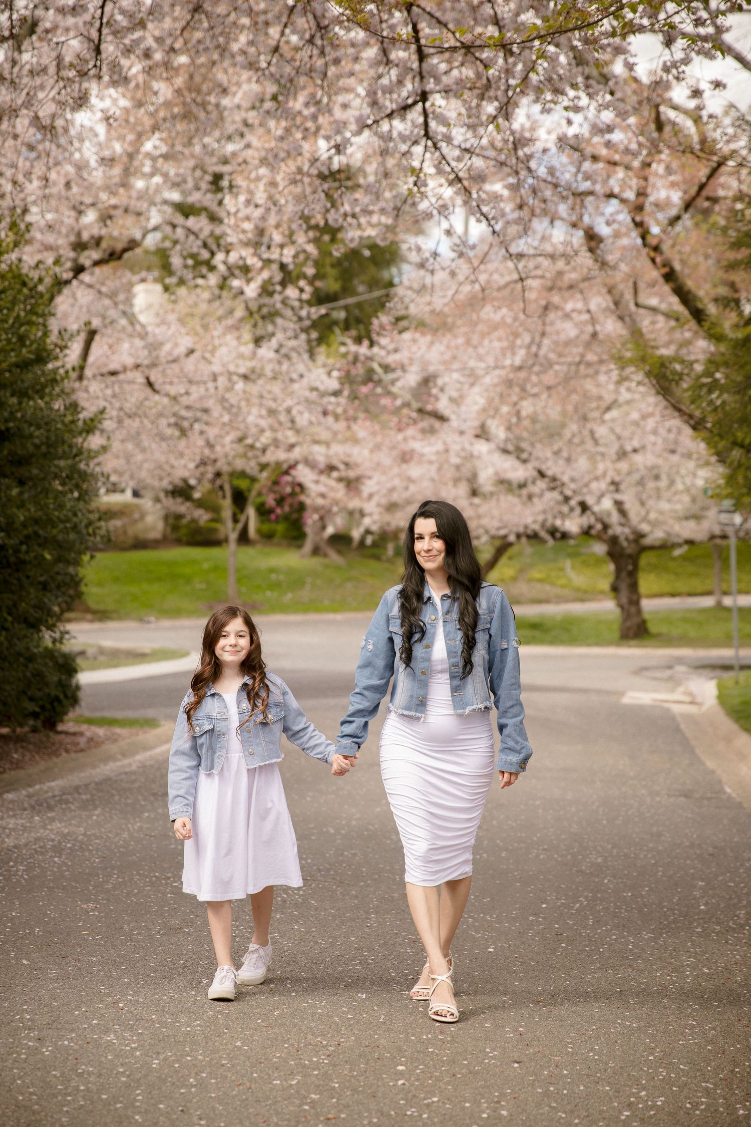 Mother and daughter in matching white dresses and denim jackets hold hands walking under cherry blossom trees.
