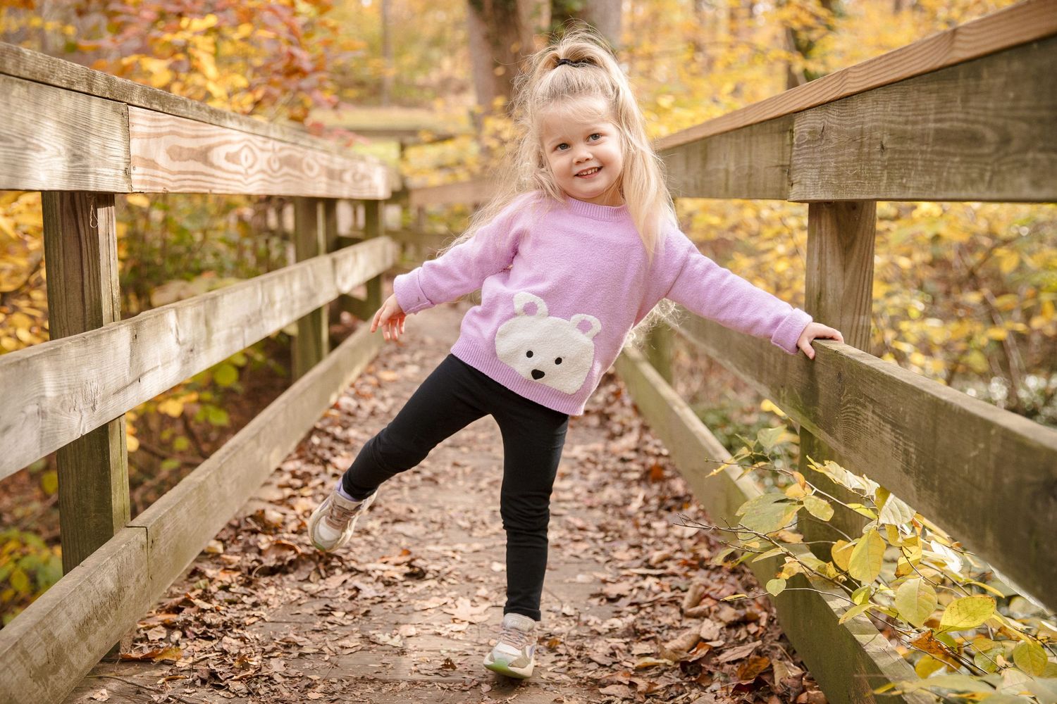 Cute blonde girl in pink bear sweater balancing on a wooden bridge in autumn forest.