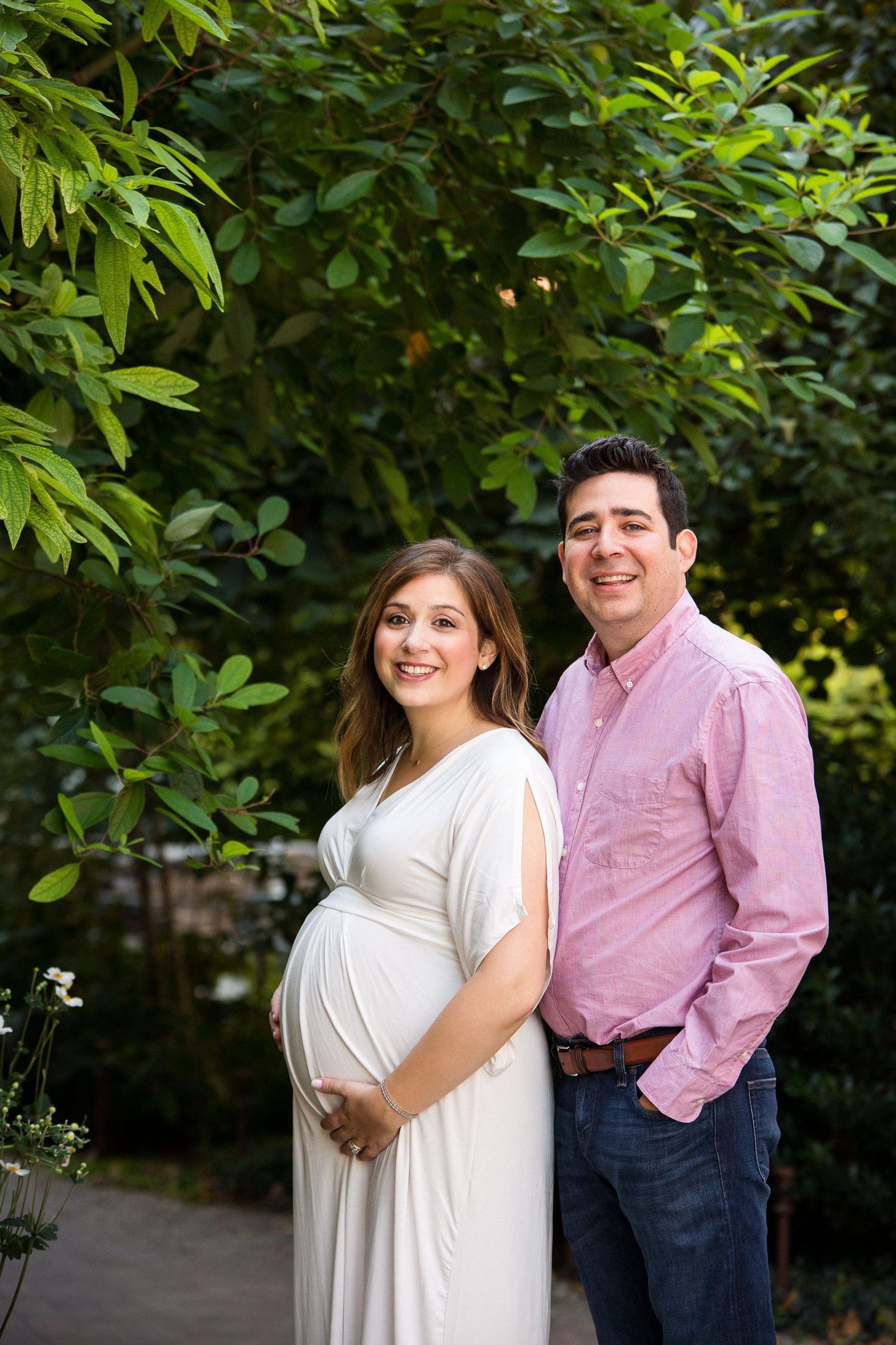 Happy expecting couple posing outdoors, pregnant woman in white dress with partner in pink shirt.
