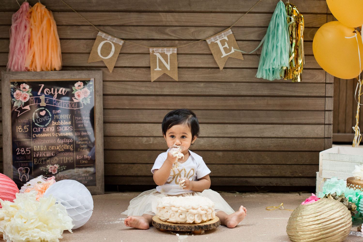 Baby girl celebrates first birthday smashing cake at ONE themed party with colorful balloons and decorations.