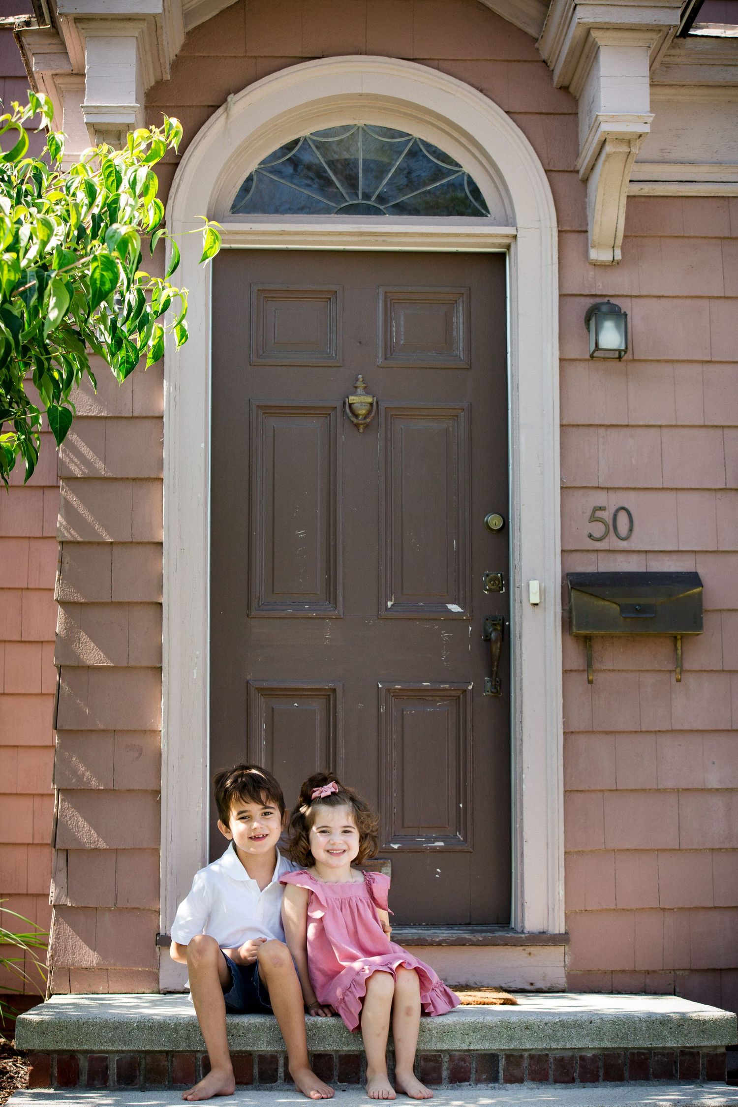 Two young children sitting barefoot on front steps of a pink house with brown door numbered 50.