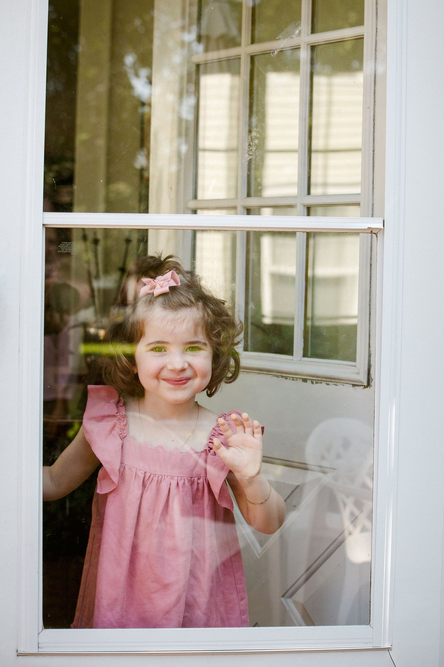 Curly-haired little girl in pink dress waving through window.