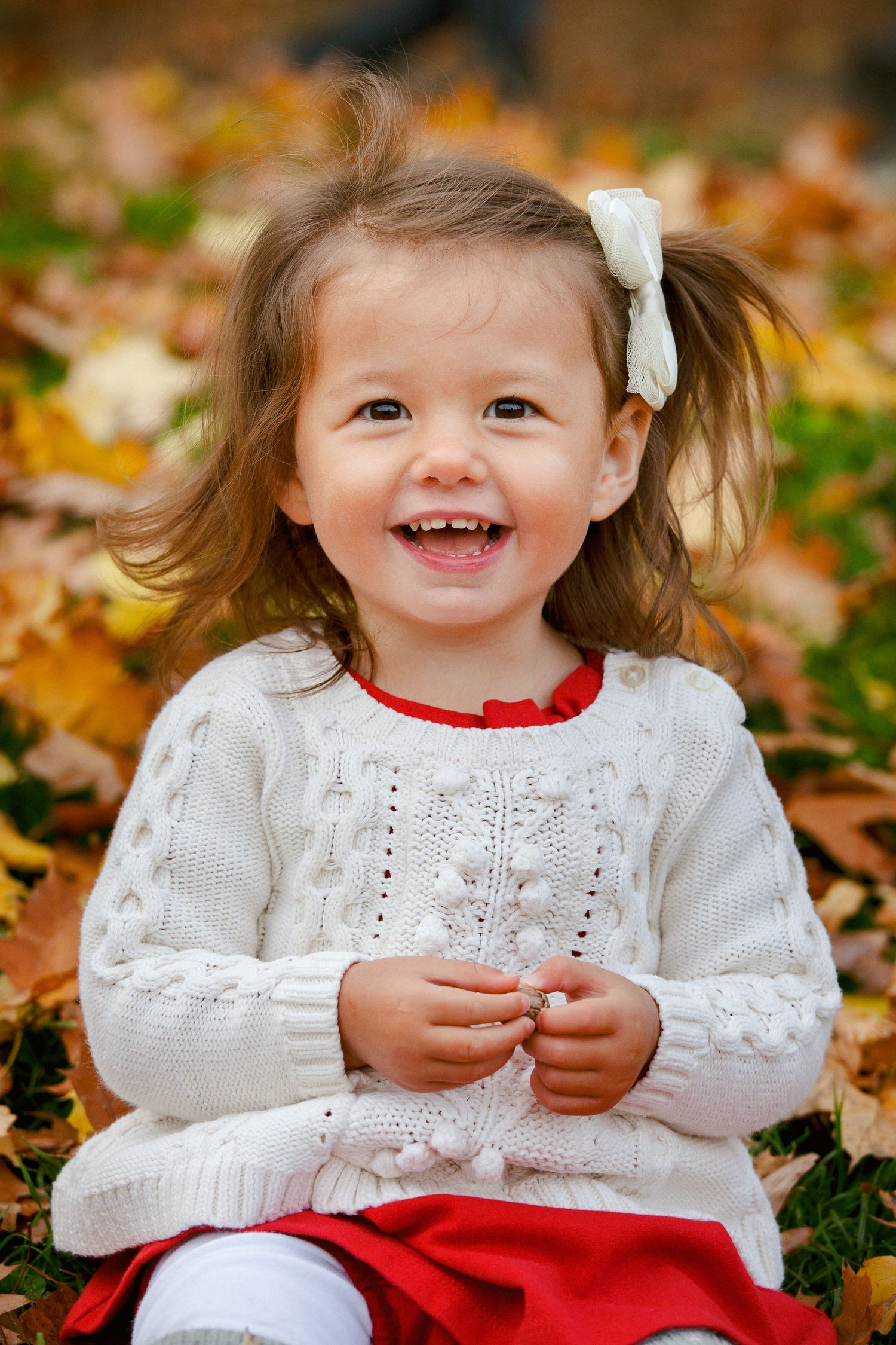 Adorable toddler girl laughing in white sweater sitting among colorful autumn leaves.