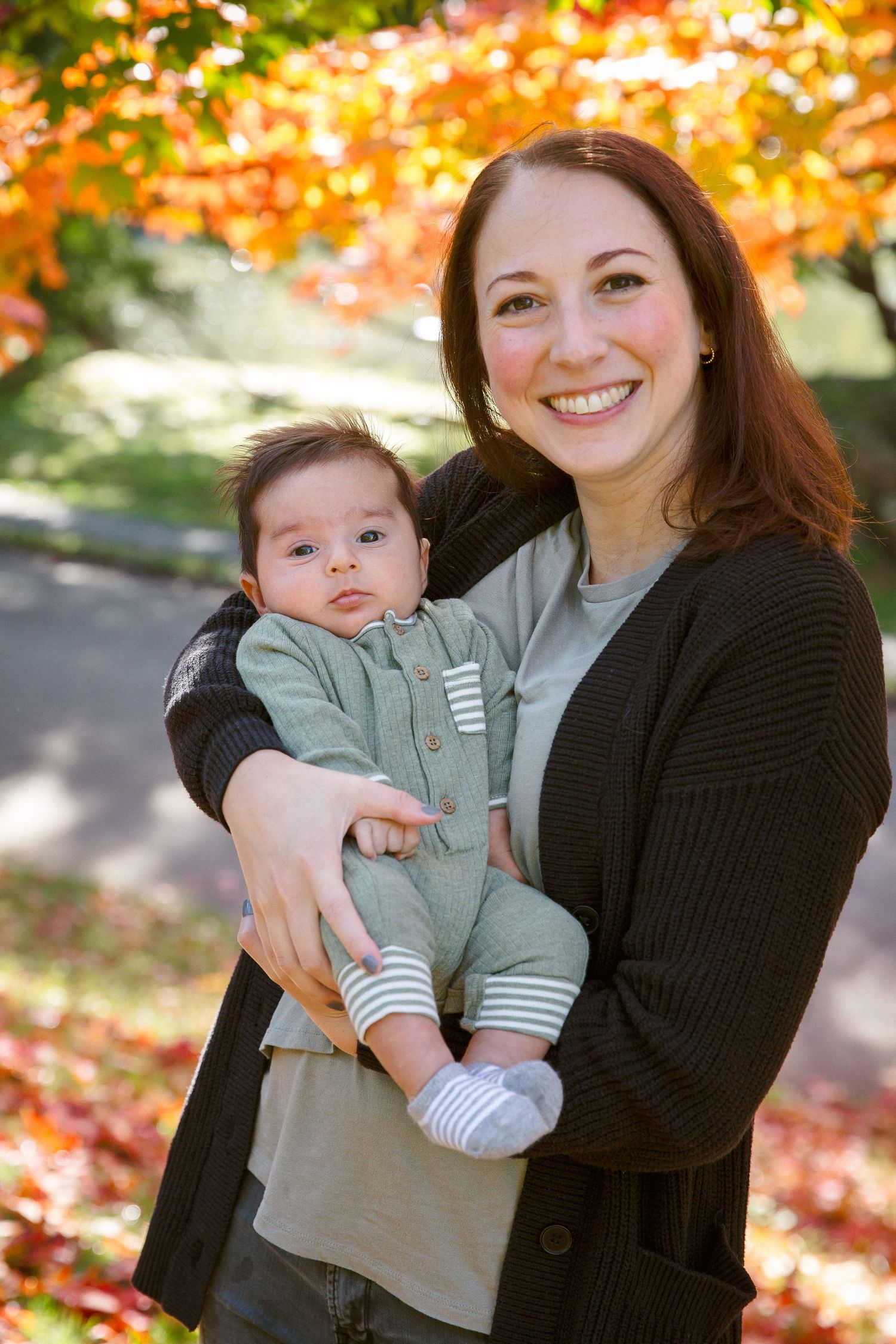 Smiling mother holding infant baby in green outfit during autumn with colorful fall foliage.