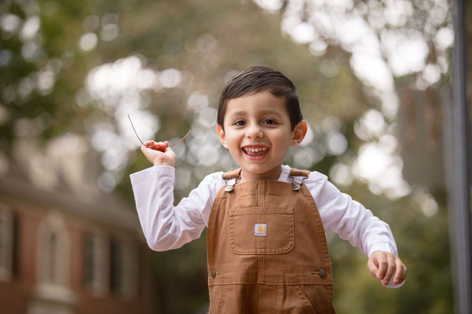 Happy toddler boy in brown Carhartt overalls smiling outdoors, holding a stick while playing outside.