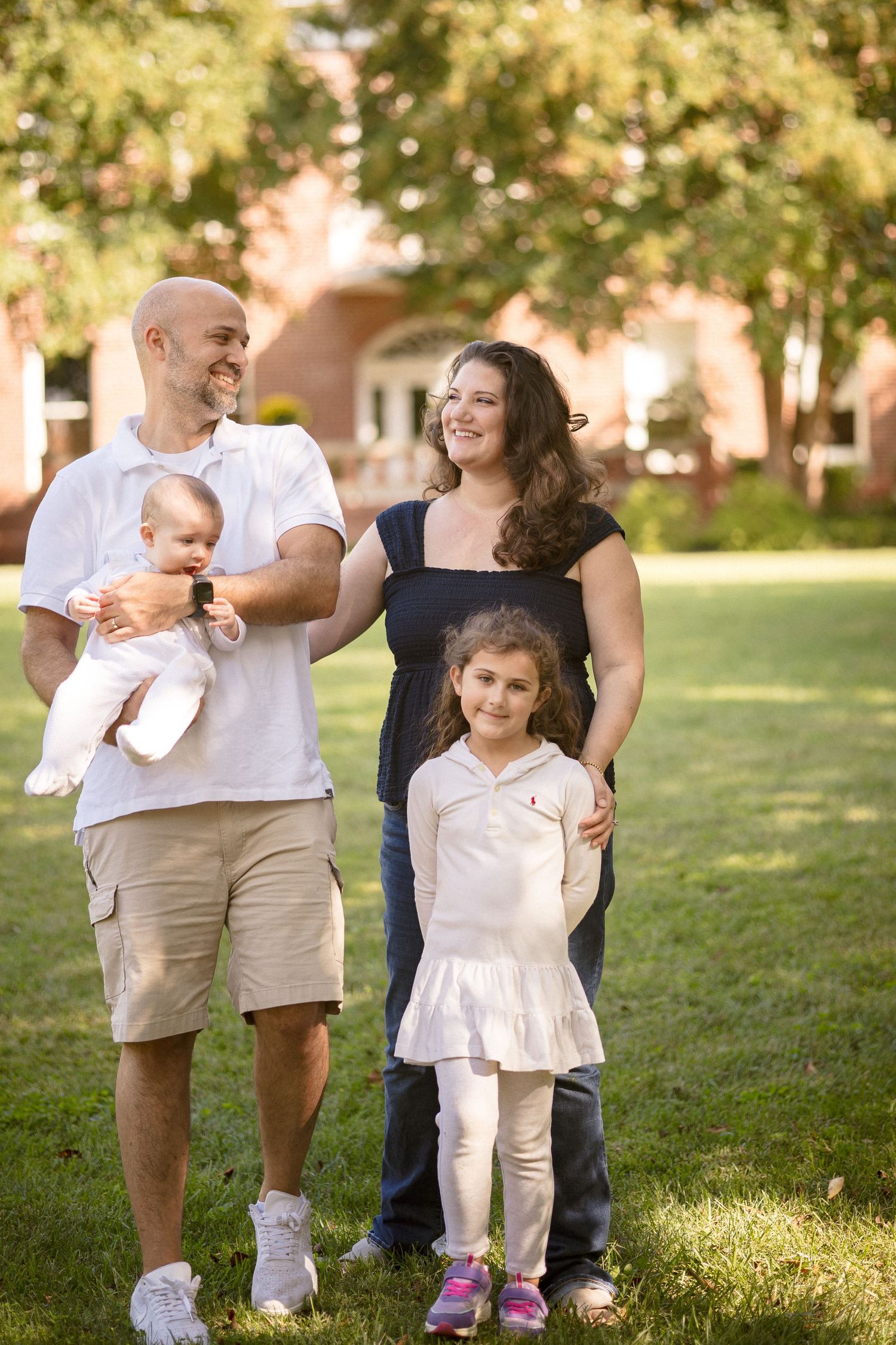 Happy family of four posing outdoors on green grass, father holding baby, mother and young daughter smiling in sunlight.