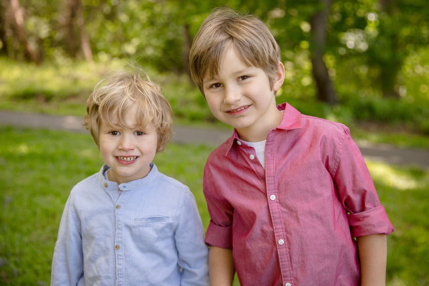 Two smiling boys in button-up shirts pose together outdoors in a lush green park setting.