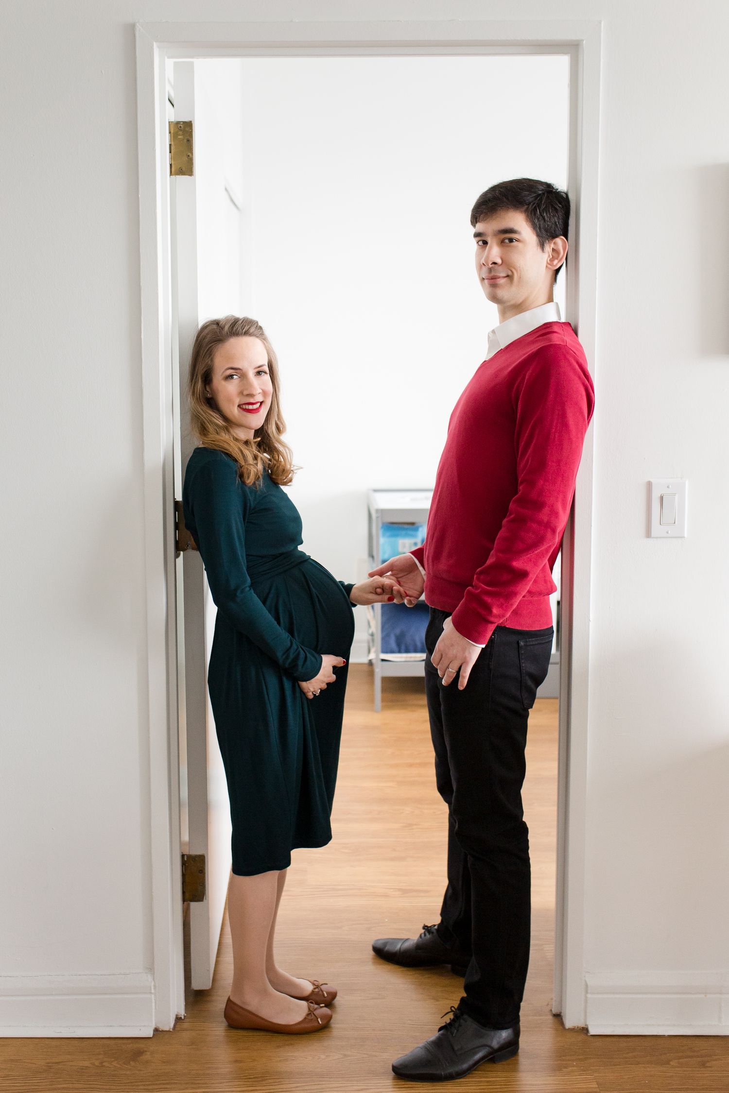 Pregnant woman in green dress and man in red sweater standing in doorway of their home.