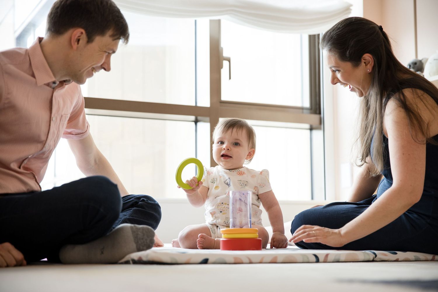 Parents sitting on floor playing with smiling baby holding colorful ring toy in bright nursery room.