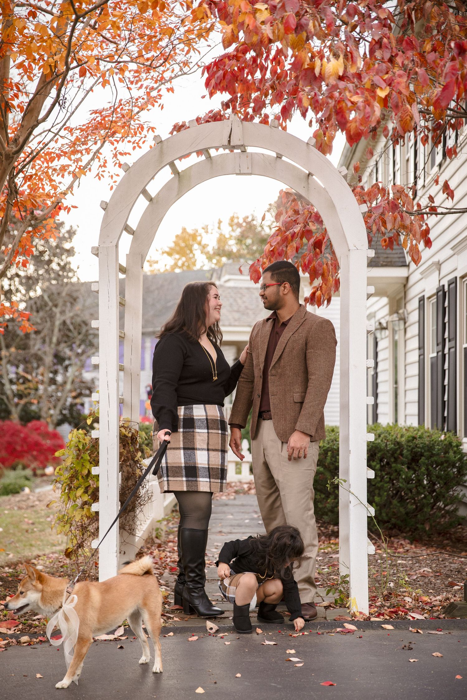 Couple standing under white garden arch with dogs amid vibrant fall foliage in a cozy suburban setting.