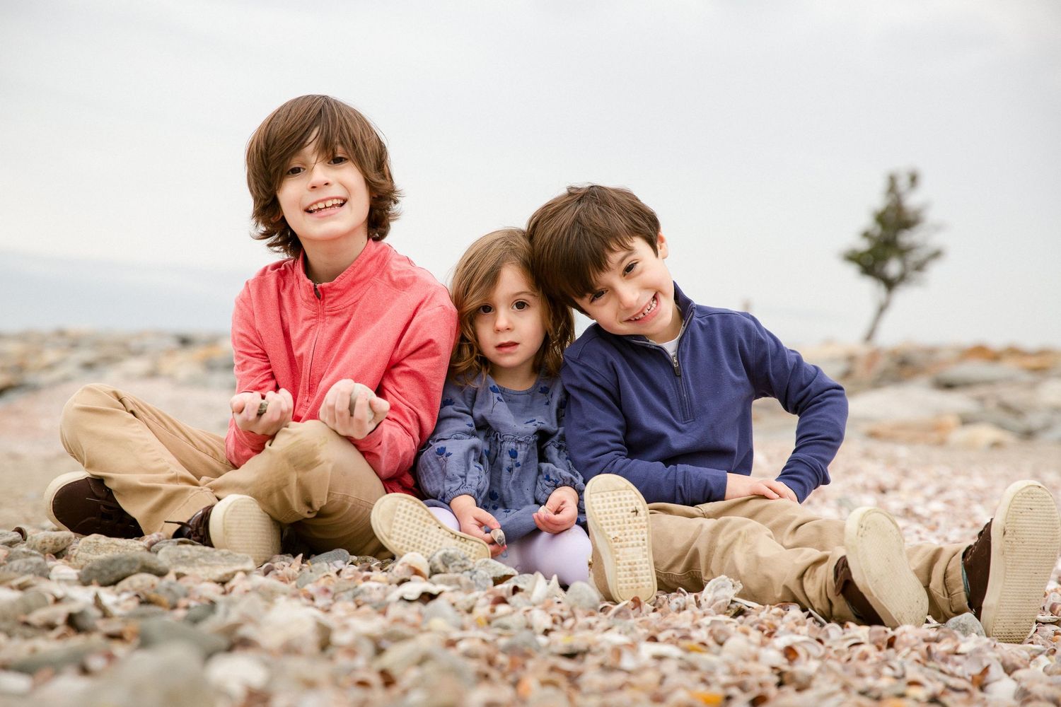 Three young siblings sit together smiling on a rocky beach on an overcast day.