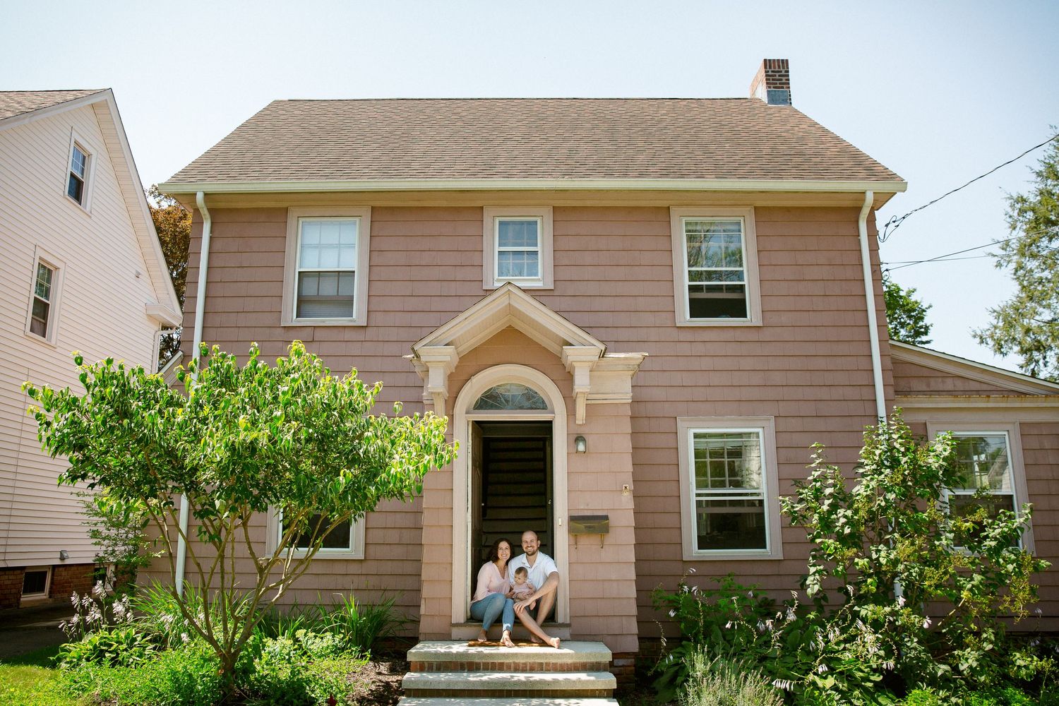 Family sitting on front steps of a charming pink colonial-style home surrounded by lush greenery.