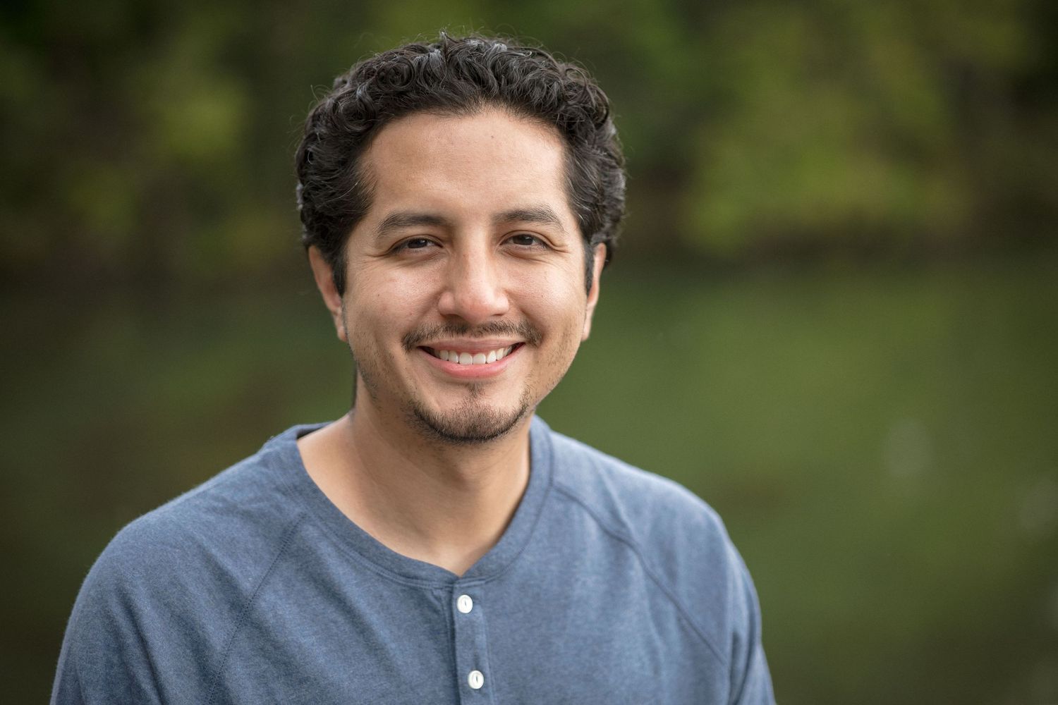 Smiling young Hispanic man in a gray henley shirt outdoors with green trees in the background.