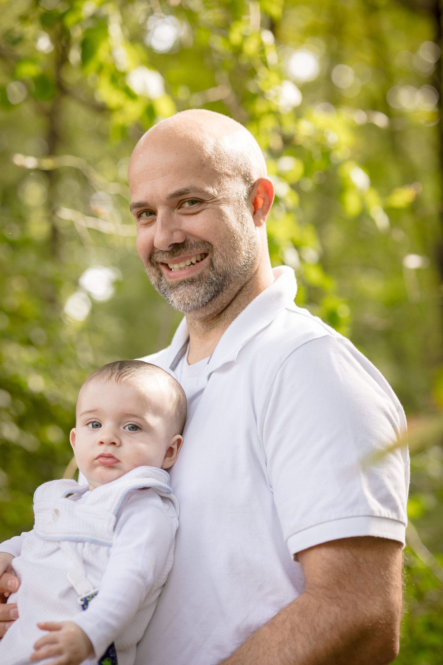 Smiling bald man with beard holding a baby outdoors in a sunny green forest setting.