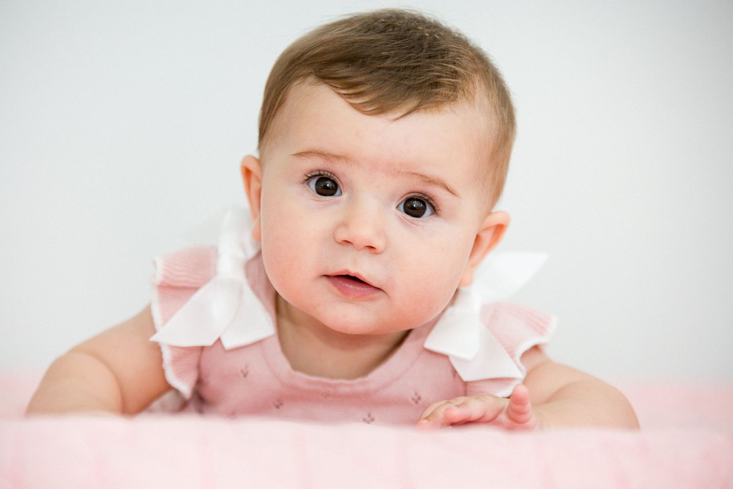 Adorable baby girl in pink ruffled dress lying on tummy with bright curious eyes.