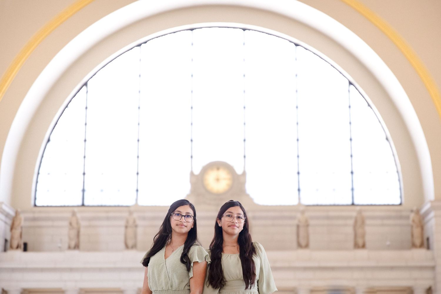 Two young women in matching sage green outfits pose inside an elegant arched building with large windows.