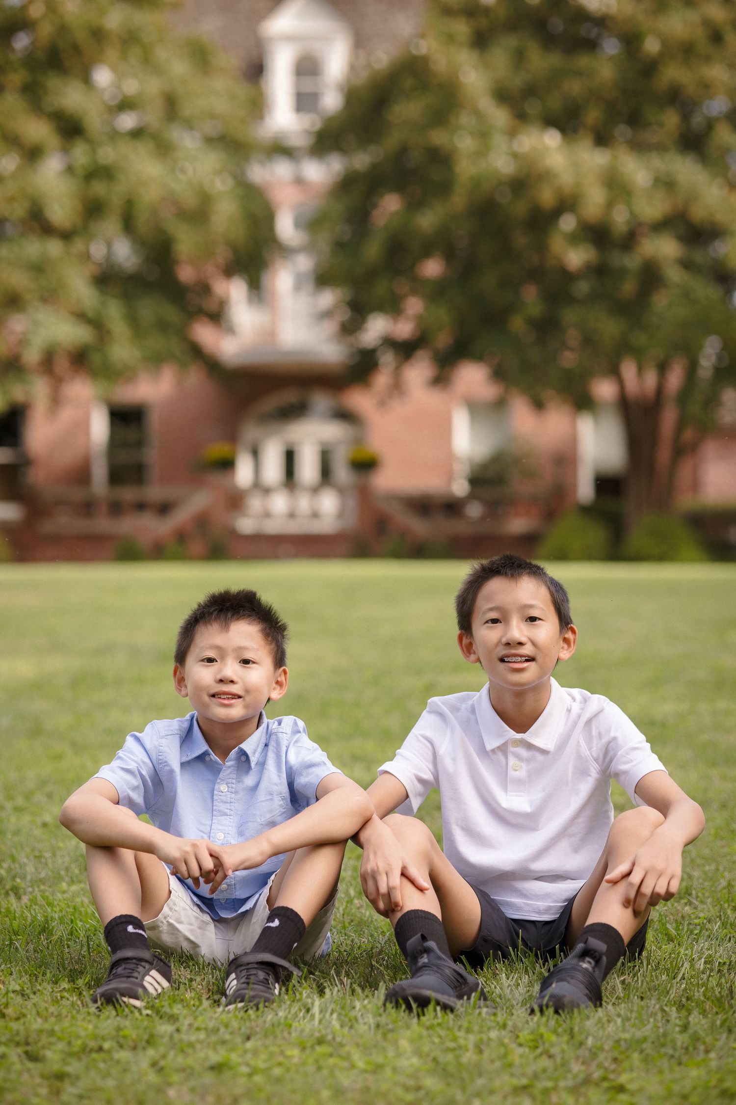 Two young Asian boys sitting on green grass outdoors, smiling, with a brick building in the background.