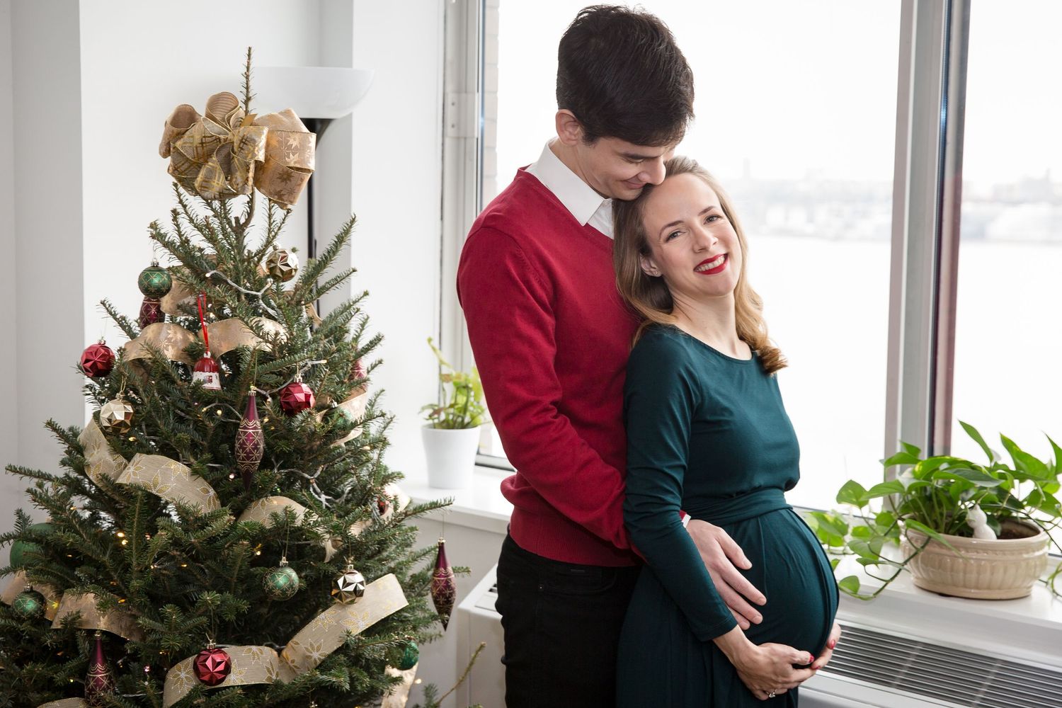 Couple posing by decorated Christmas tree, pregnant woman in green dress, man in red sweater, near bright window.