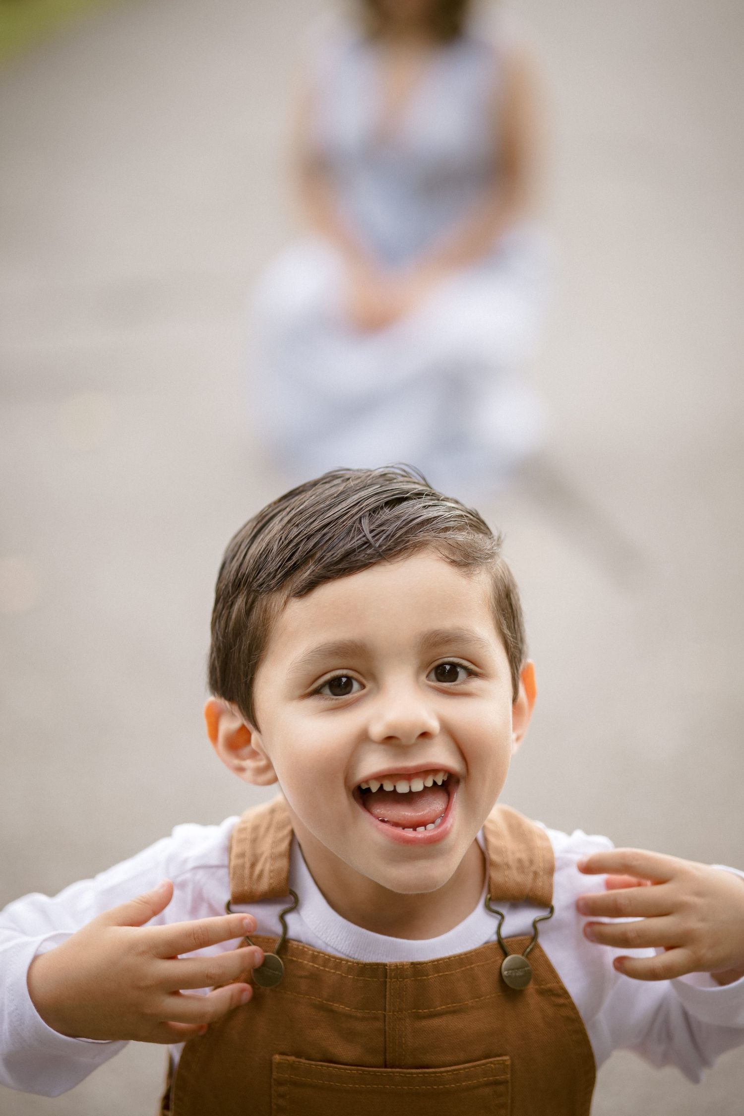 A happy young boy in brown overalls and white shirt makes a funny face outdoors.