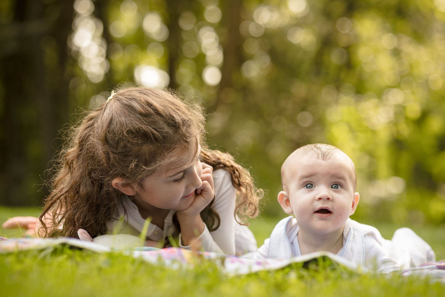 Young girl and baby lying on grass in a sunny park, looking at each other.