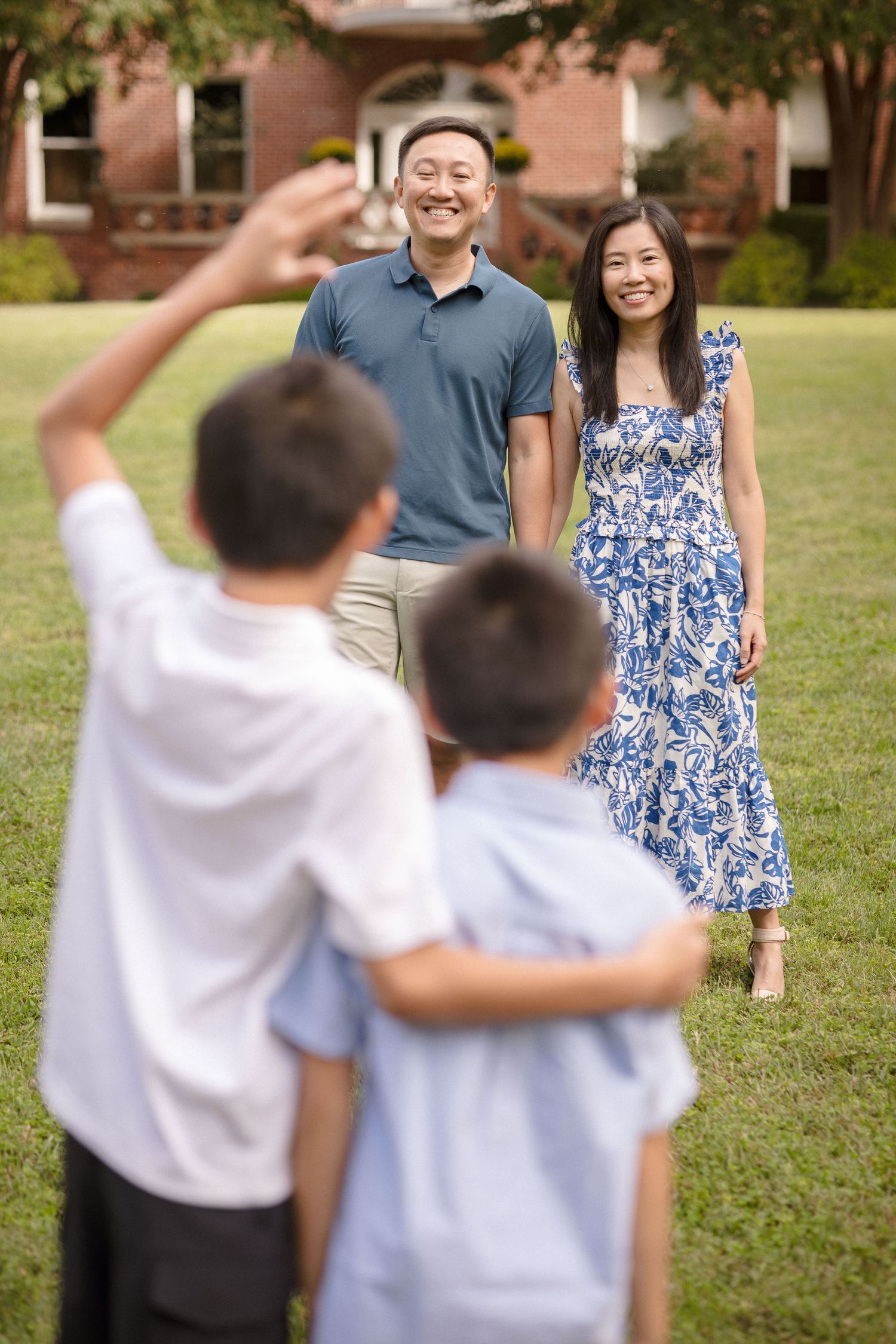 Happy Asian family with two boys greeting their smiling parents on a sunny lawn near a brick building.
