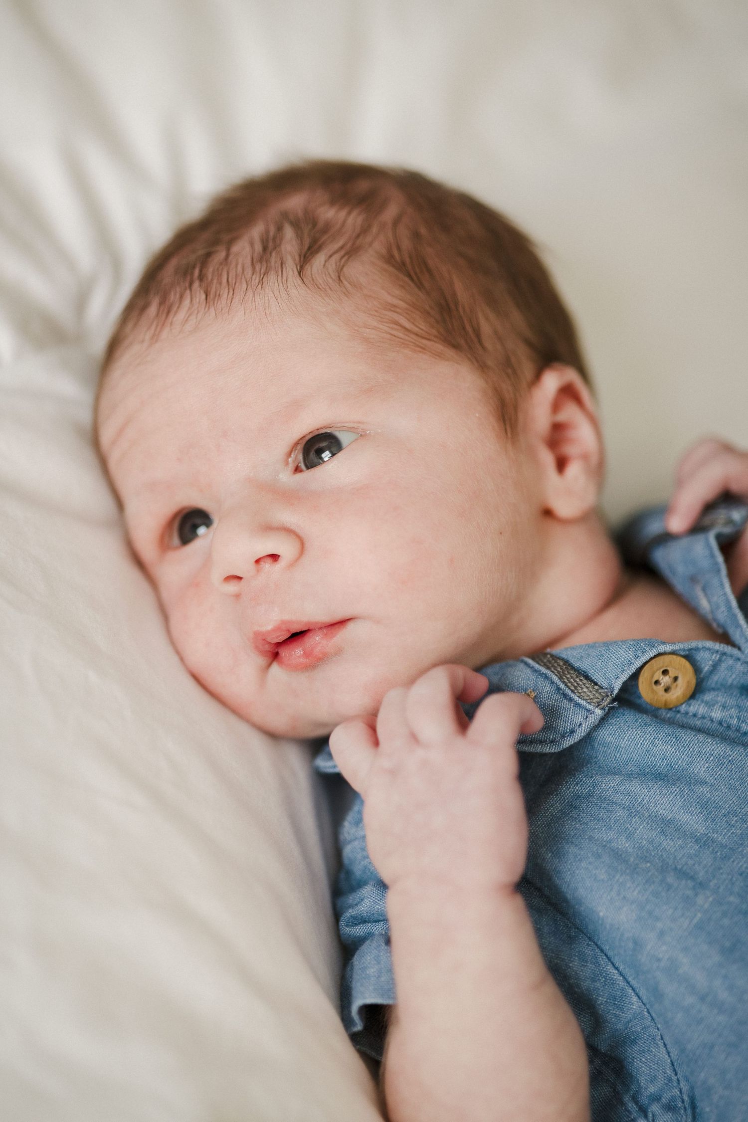 Newborn baby in denim romper lying on white blanket looking up with curious expression.