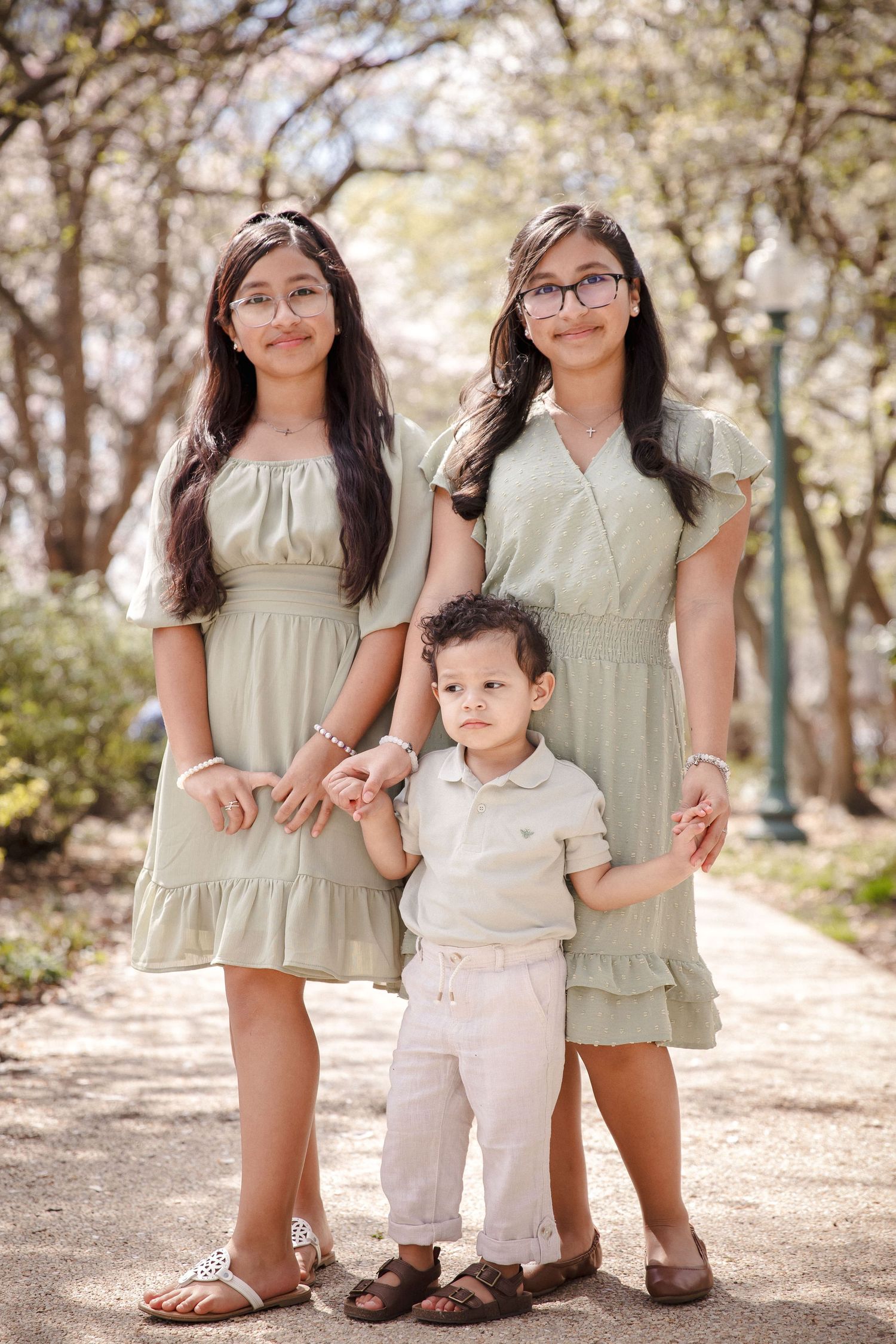 Two teenage sisters in sage dresses pose with their little brother in a park.