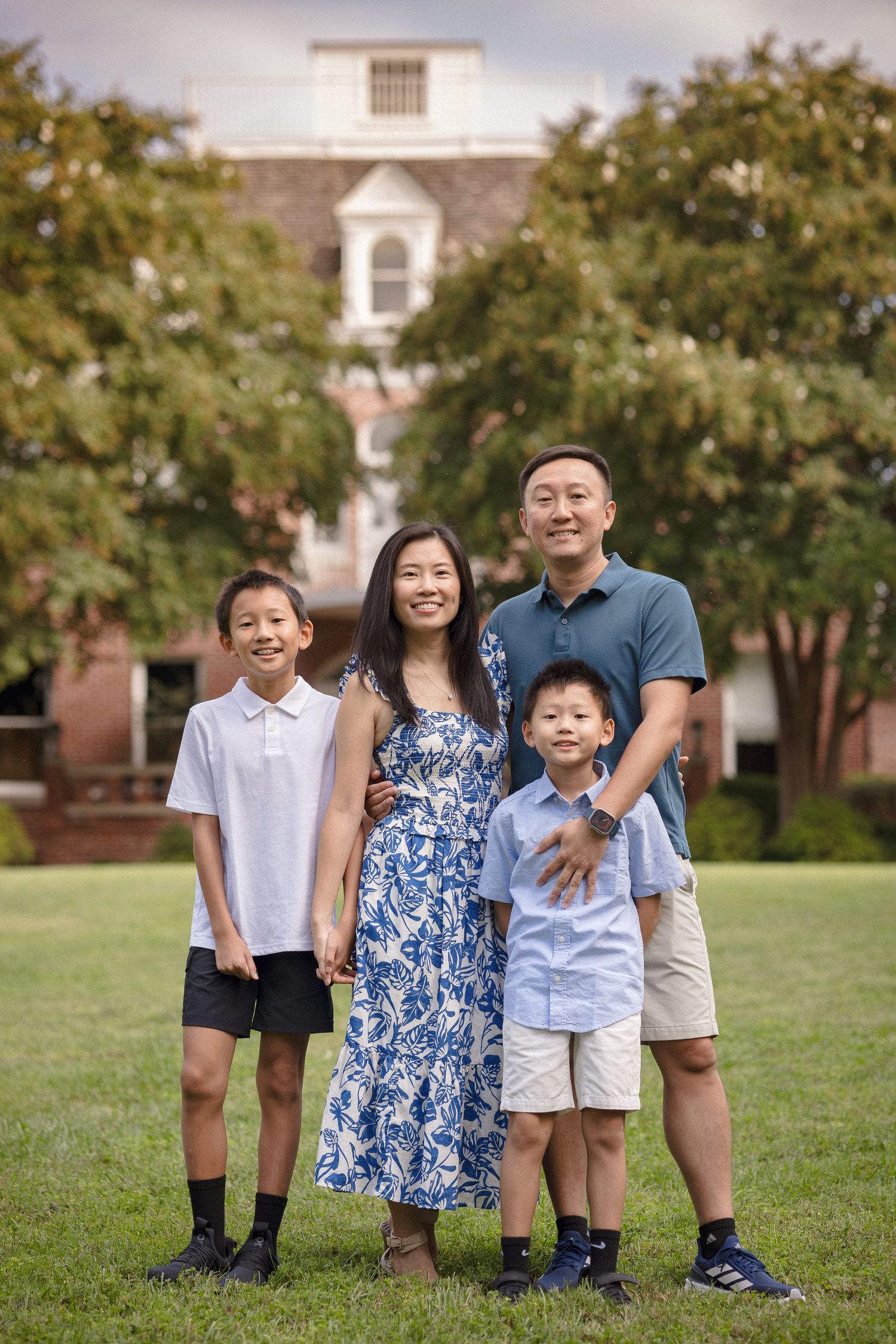 Asian family of four posing outdoors on green lawn with brick building and trees in background.