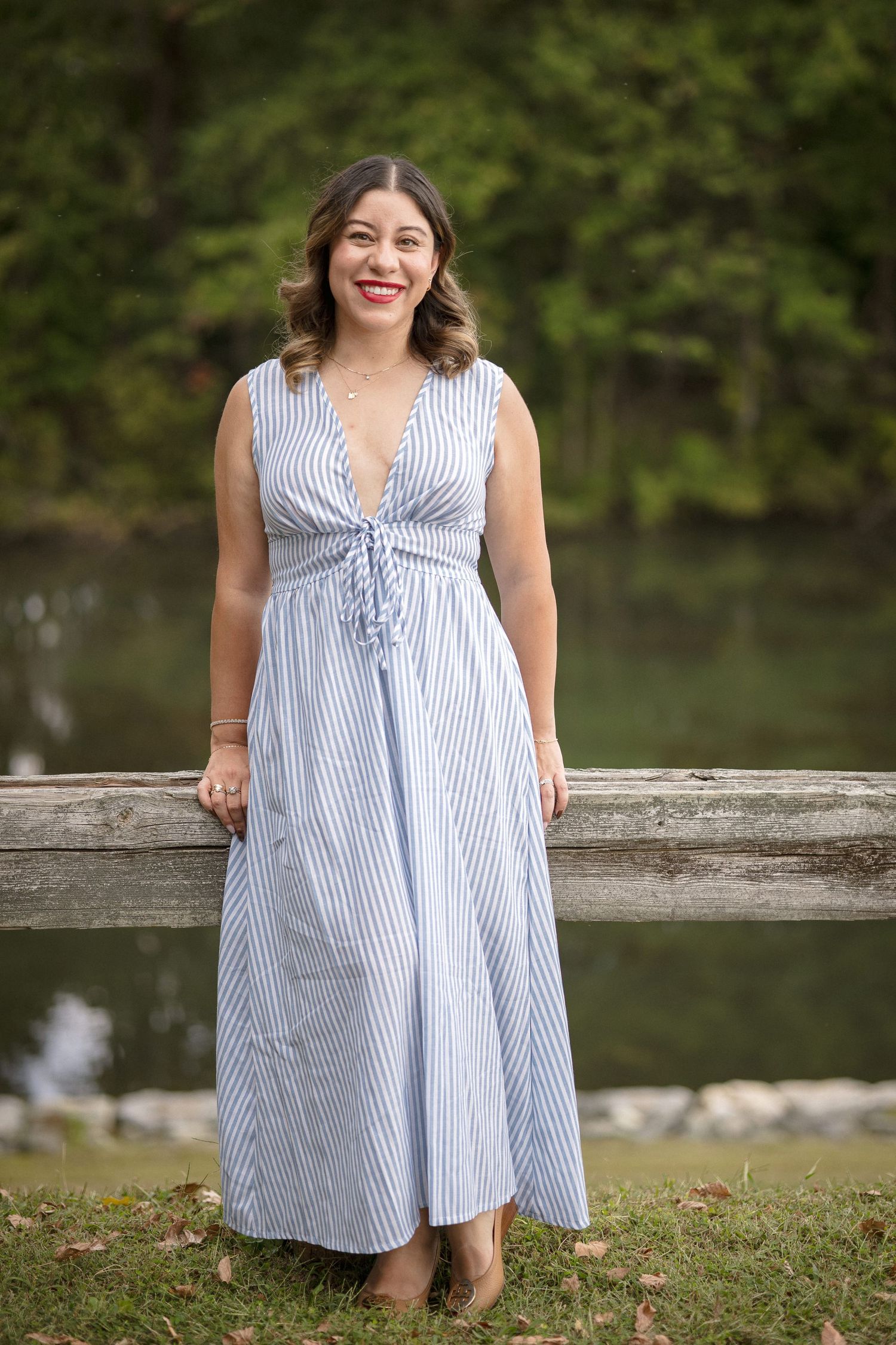 Woman in blue striped maxi dress smiling outdoors near wooden fence and pond with green trees background.