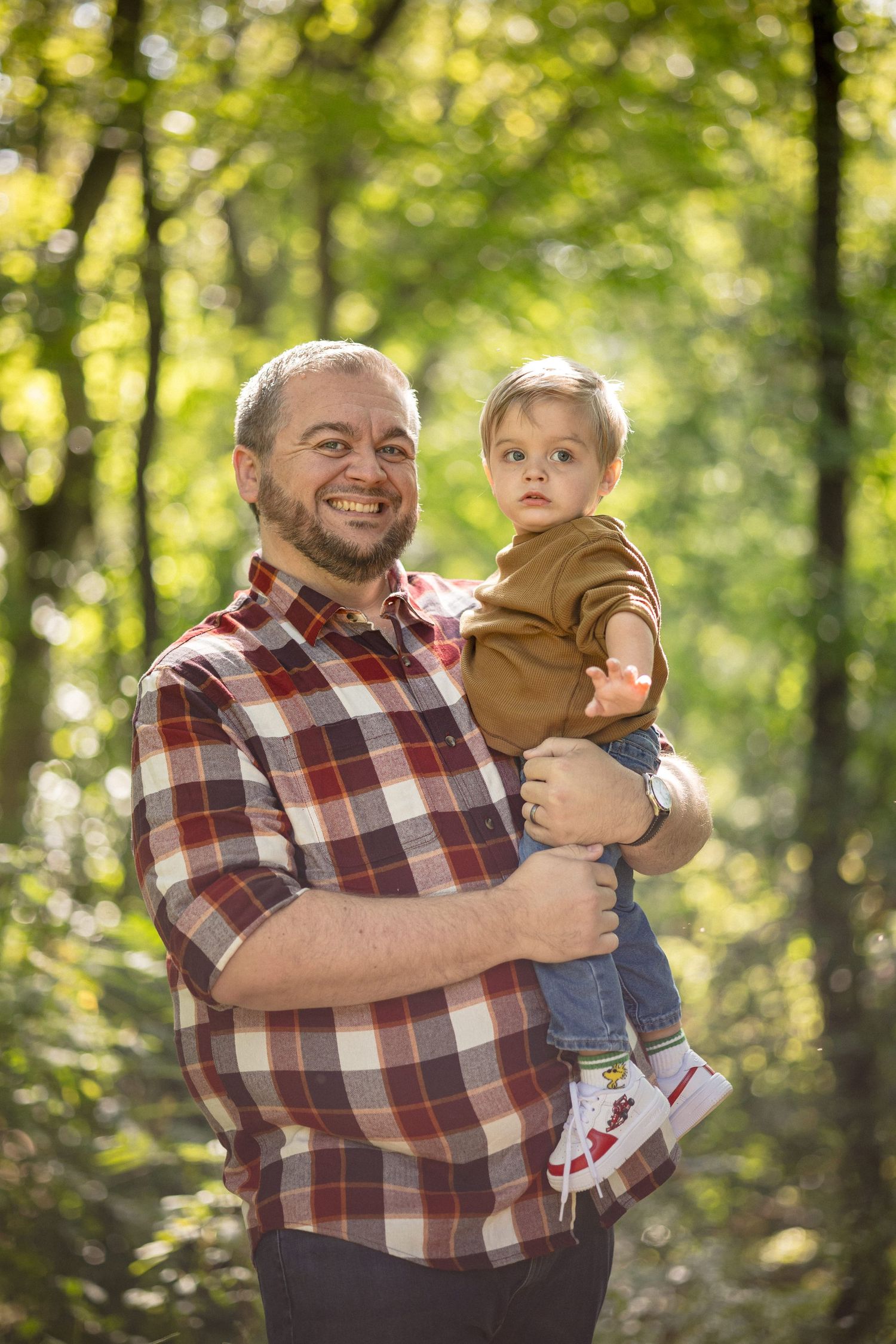 A smiling father in a plaid shirt holds his toddler in a sunlit forest setting.